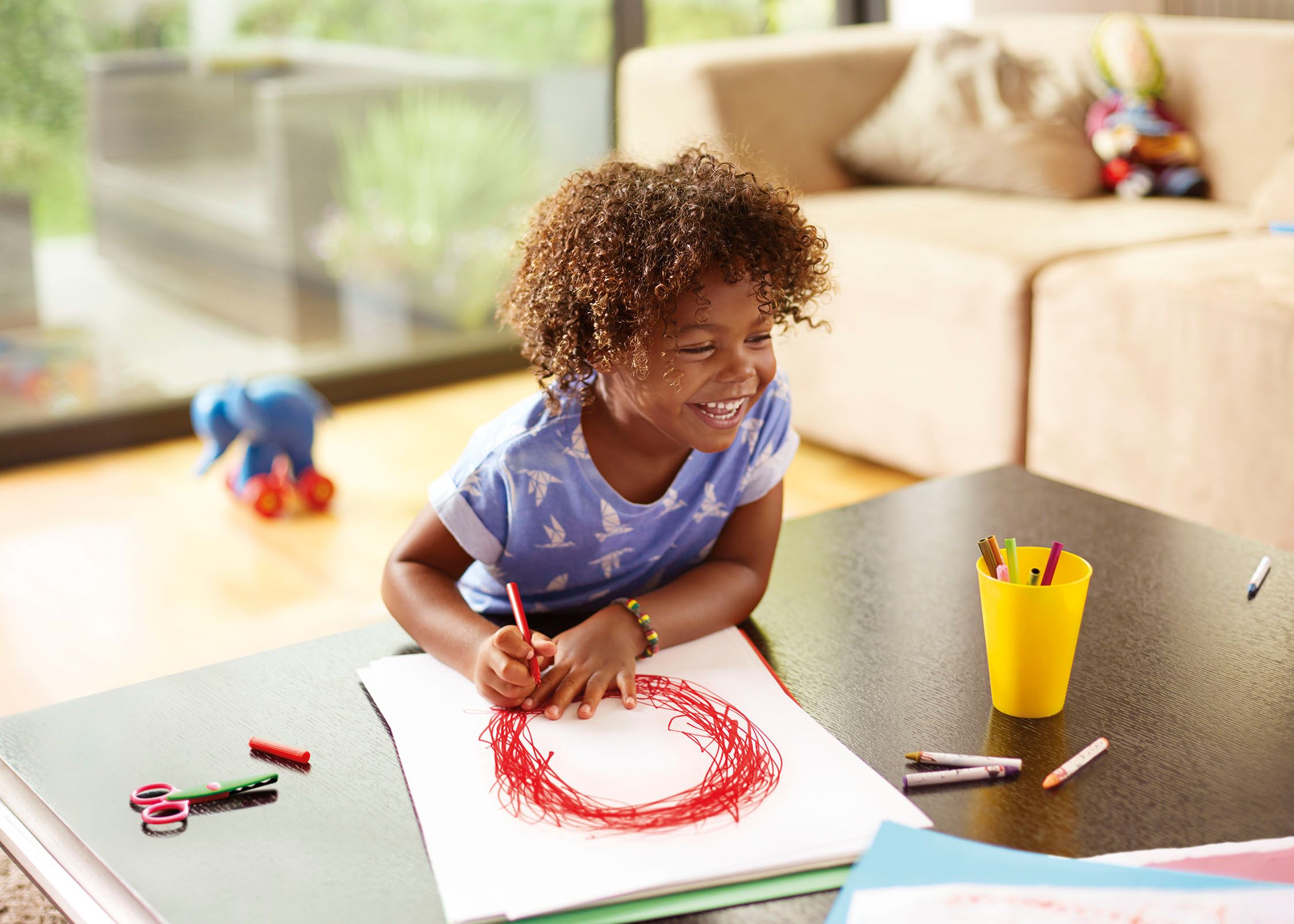 A young girl with curly hair is sitting at a table, drawing with red crayons on a white sheet of paper.