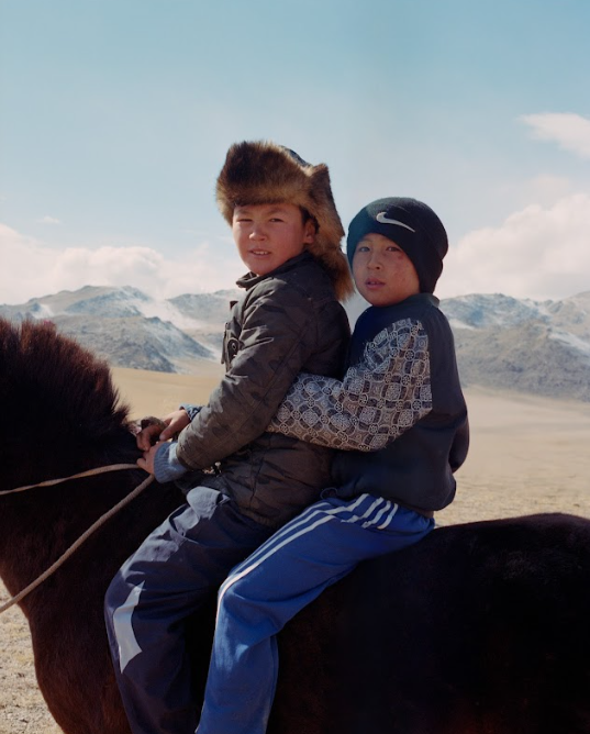Two children, one wearing a fur hat, riding a horse in a mountainous landscape.