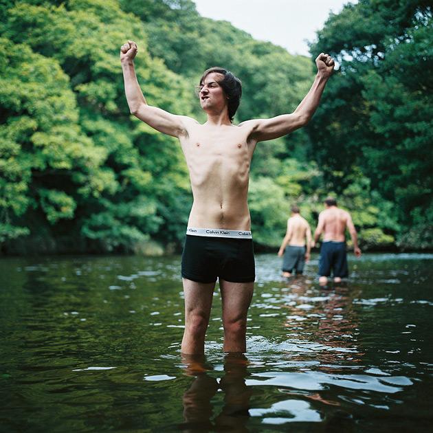A young man in a wetsuit, standing in a river, with his arms raised in triumph.