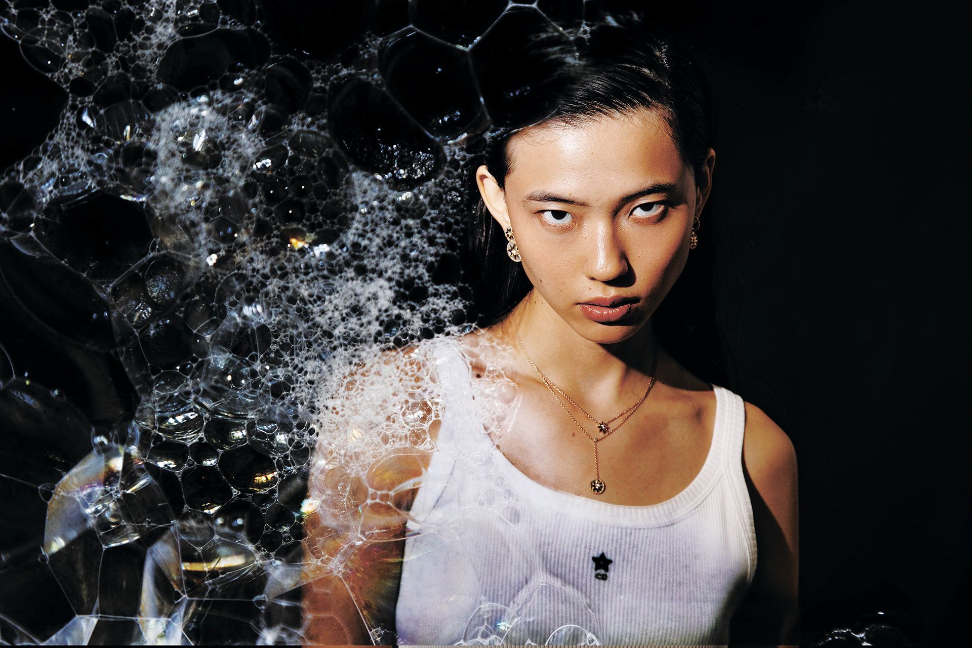 A young woman with dark hair and makeup, wearing a white tank top and a gold necklace with a pendant, in front of a dark background with a subtle, abstract, bubble-like pattern.