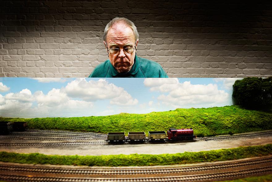 An older man with a serious expression, wearing a green sweater, standing in front of a white brick wall.