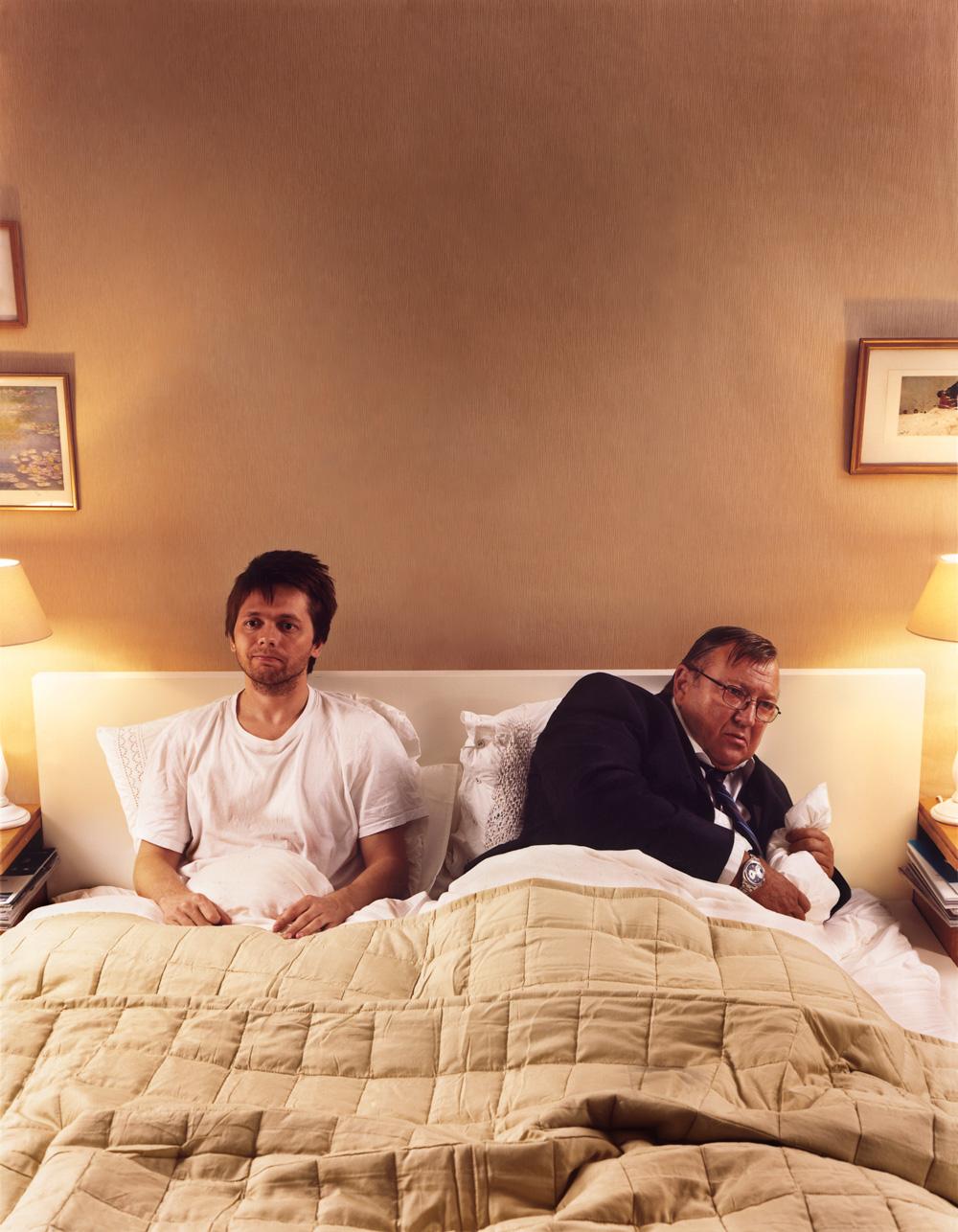 A man in a white t-shirt and a man in a suit are lying in bed, with the man in the suit covering his face.