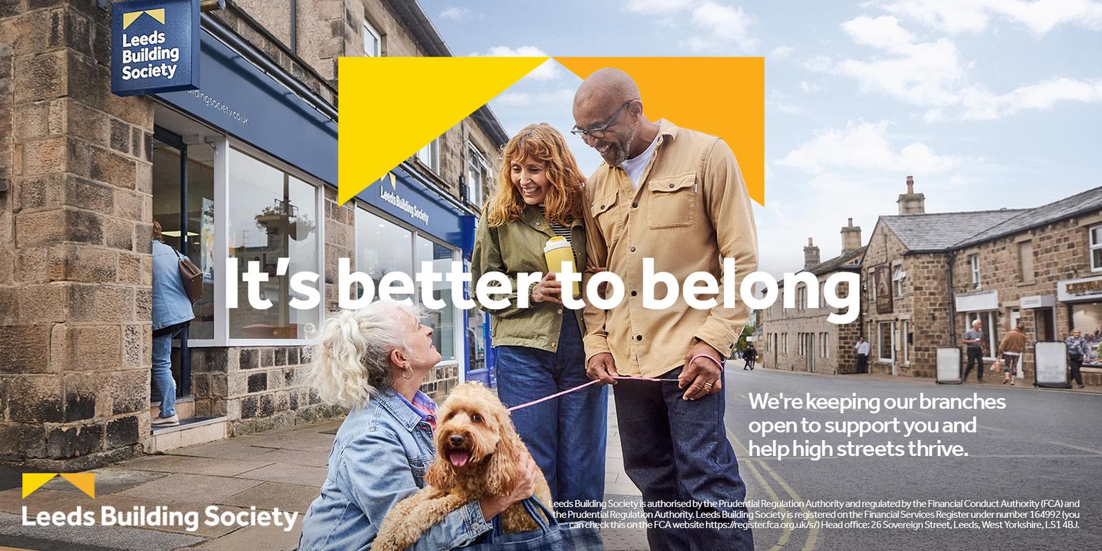 A stone building with a blue sign reading "Leeds Building Society" and a yellow triangle in the corner. Two people, one with a dog, are in front of the building, while a person.