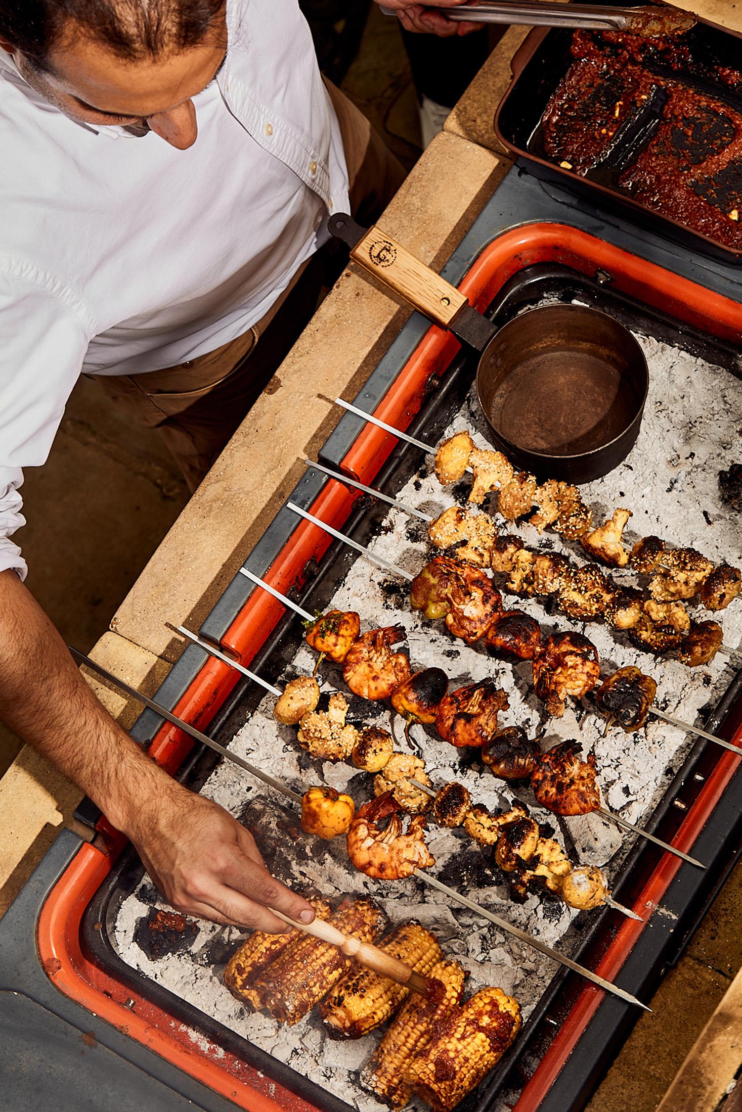 A man in a white shirt is preparing skewers of meat and vegetables on a grill.