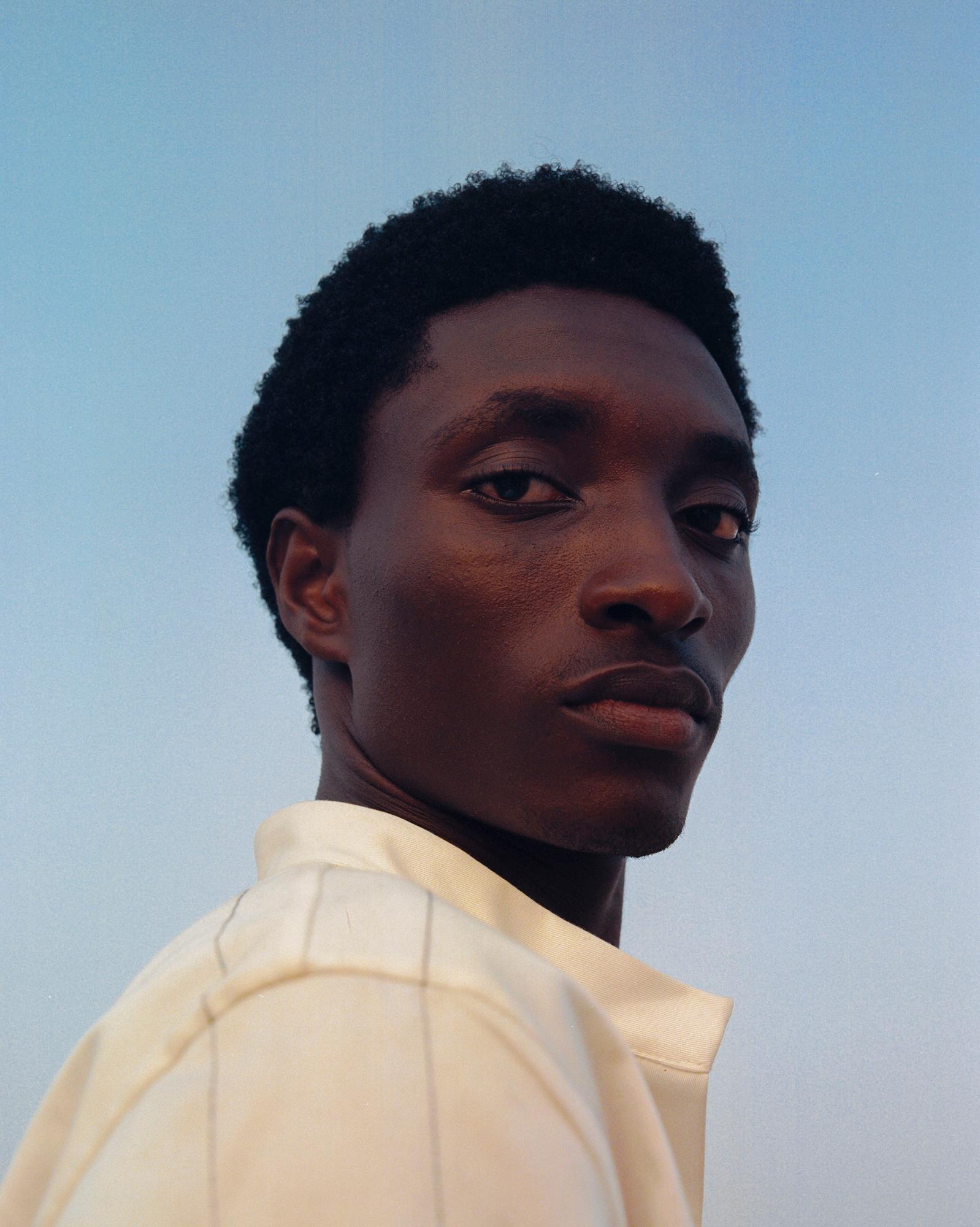 A young man with a short, dark afro and a small, dark beard, wearing a white, long-sleeved, button-down shirt with a subtle, horizontal, dark stripe pattern.