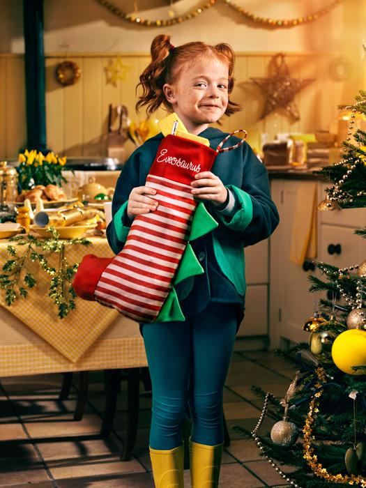 A young girl holding a large, red, striped Christmas stocking with her name 'Evensaurel' embroidered on it, standing in a festive kitchen with a table set for a holiday meal.