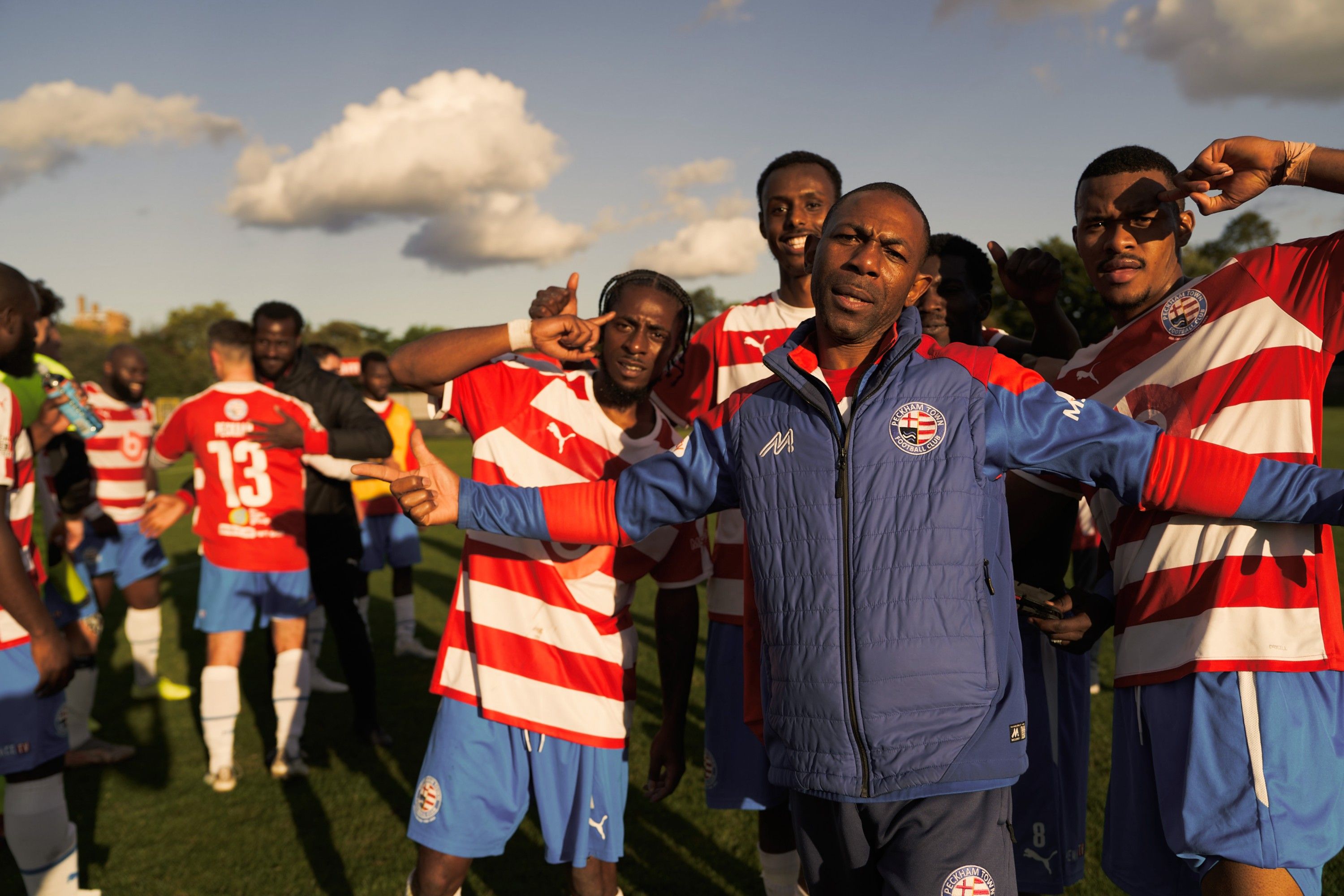 A group of men in football jerseys and one man in a blue jacket standing on a football field, with some of them making hand gestures.