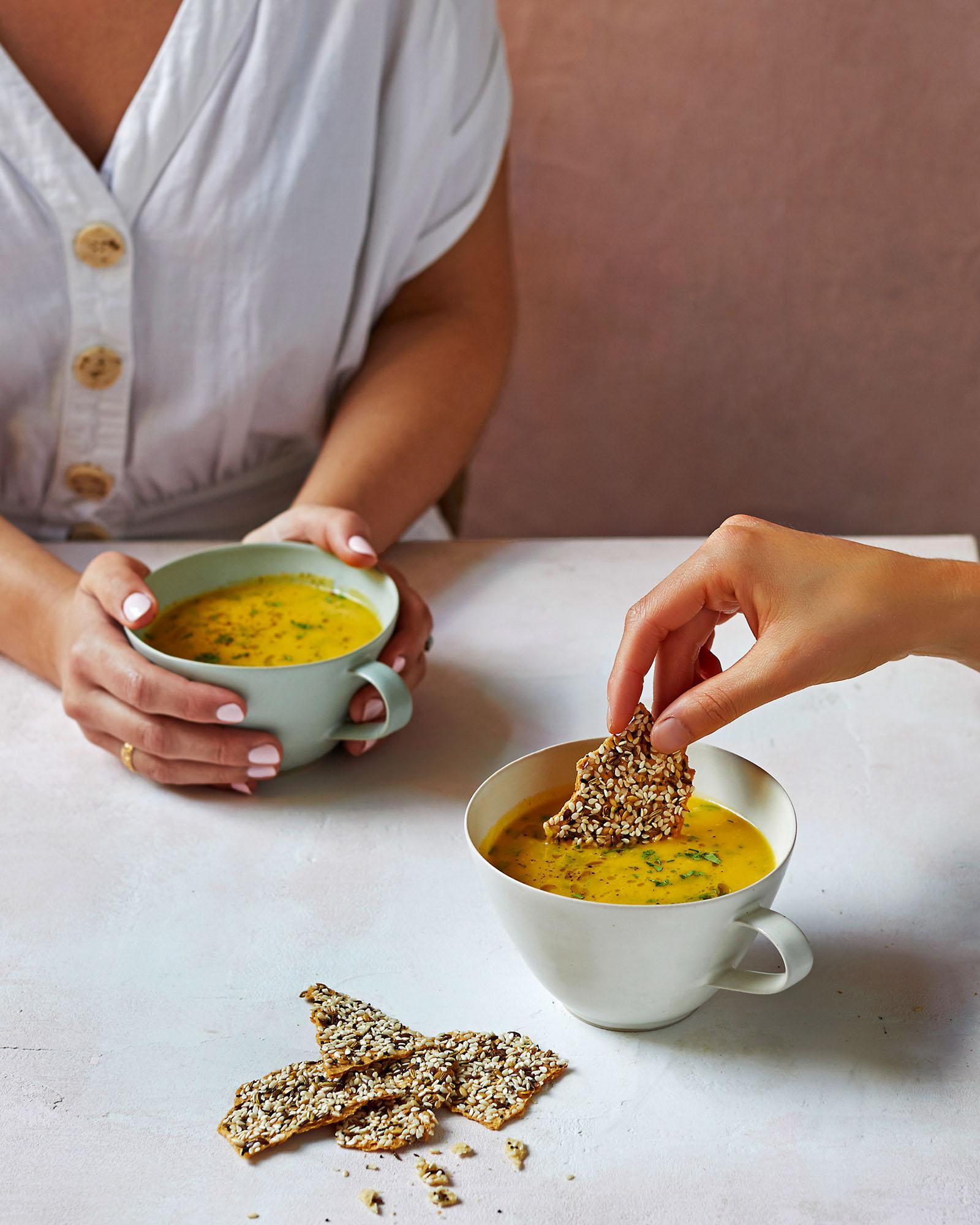 A woman in a white blouse and a man in a white shirt are holding cups of soup and dipping a cracker into it.