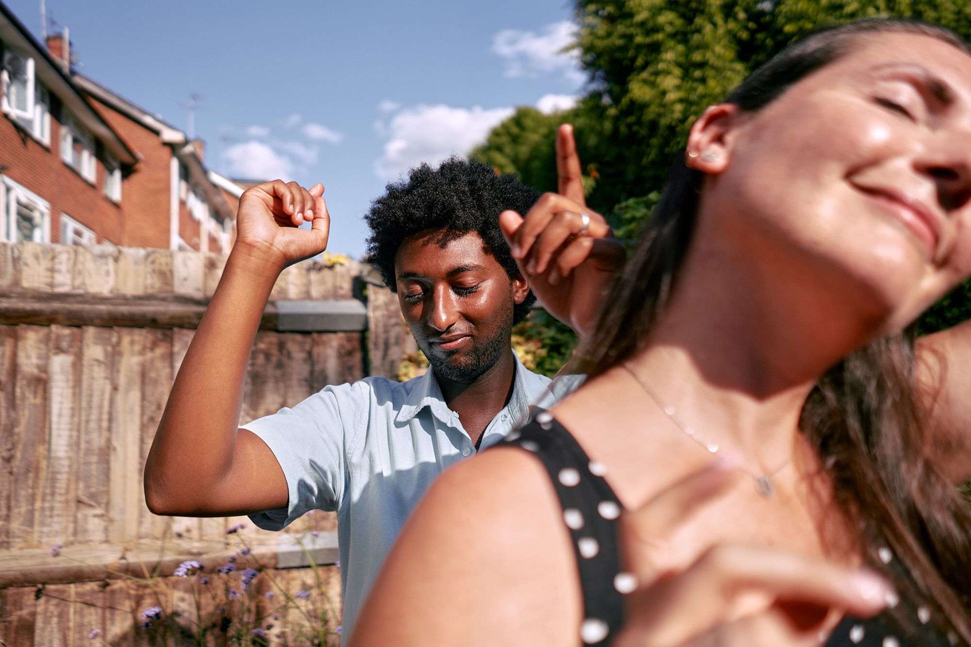 A man with a beard and a woman with long hair are dancing in a backyard, surrounded by a wooden fence and a few trees, with a blue sky and a few clouds in the background.