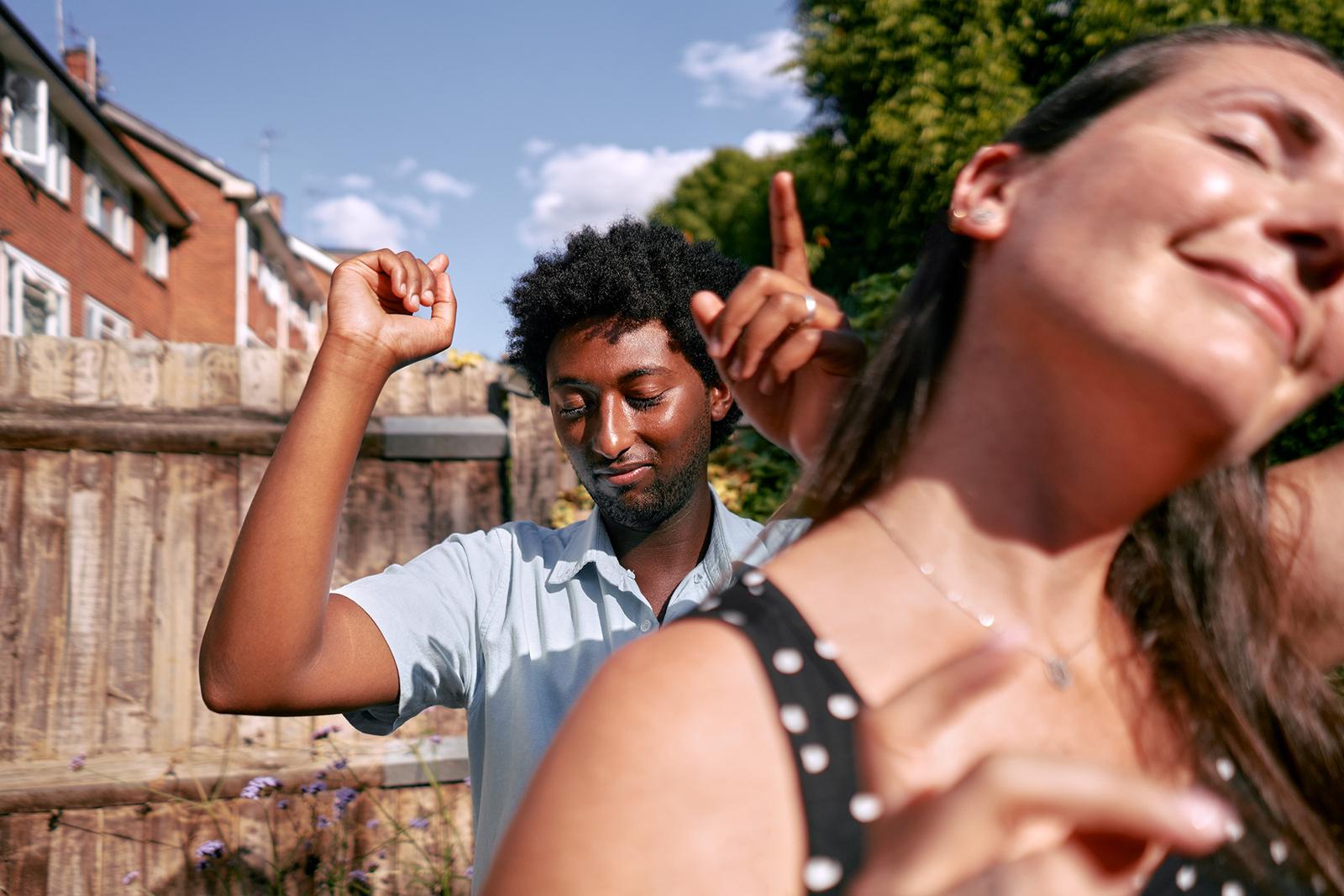 A man with a beard and a woman with long hair are dancing in a backyard, surrounded by a wooden fence and a few trees, with a blue sky and a few clouds in the background.