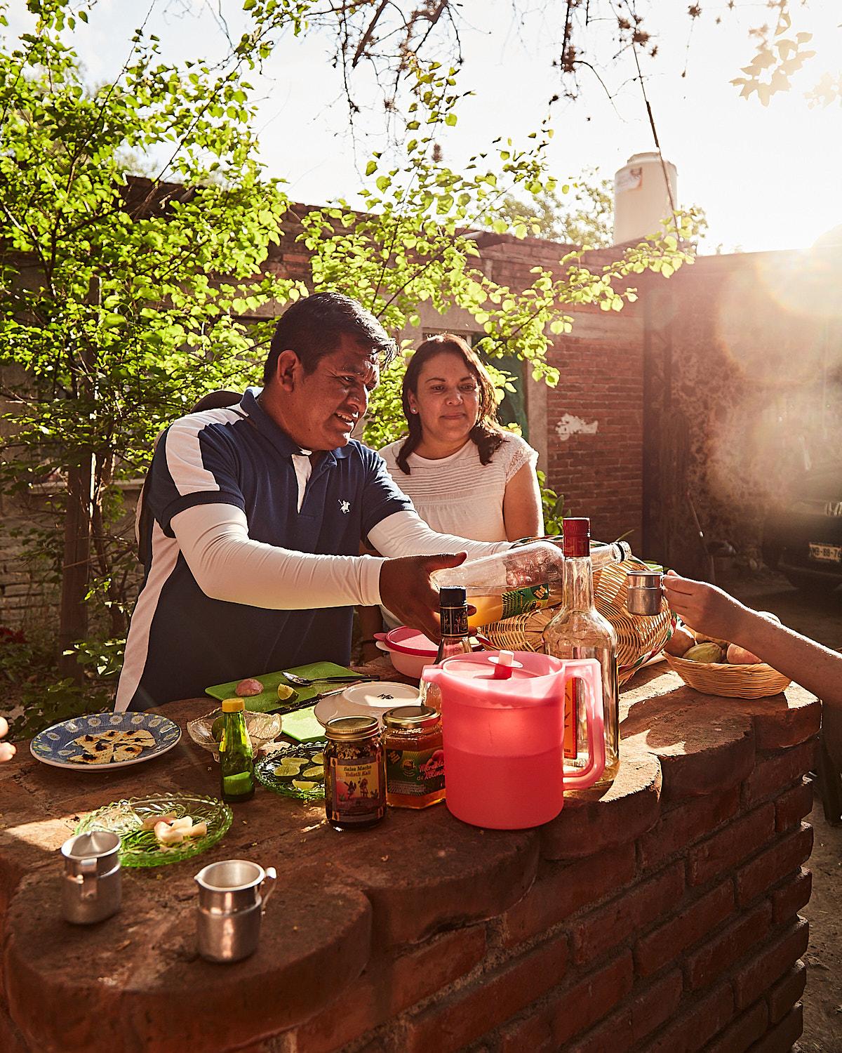 A man in a blue and white striped polo shirt is pouring a drink into a glass for a woman in a white top, both standing behind a brick bar in a garden setting.