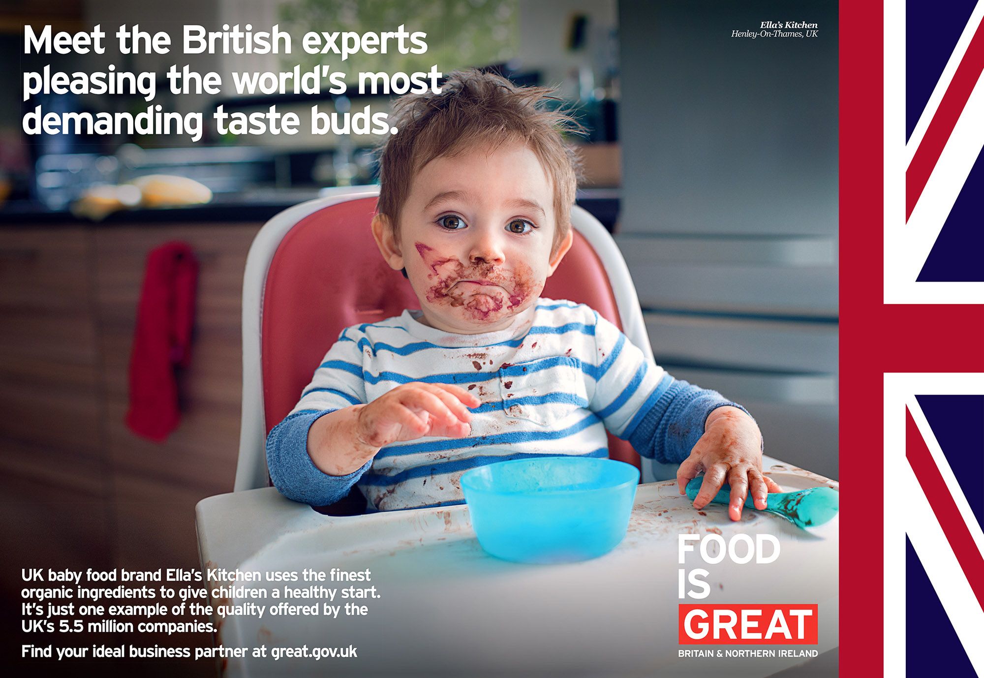 A toddler sits in a high chair, covered in chocolate and food, with a messy face and a spoon in hand, in a kitchen.