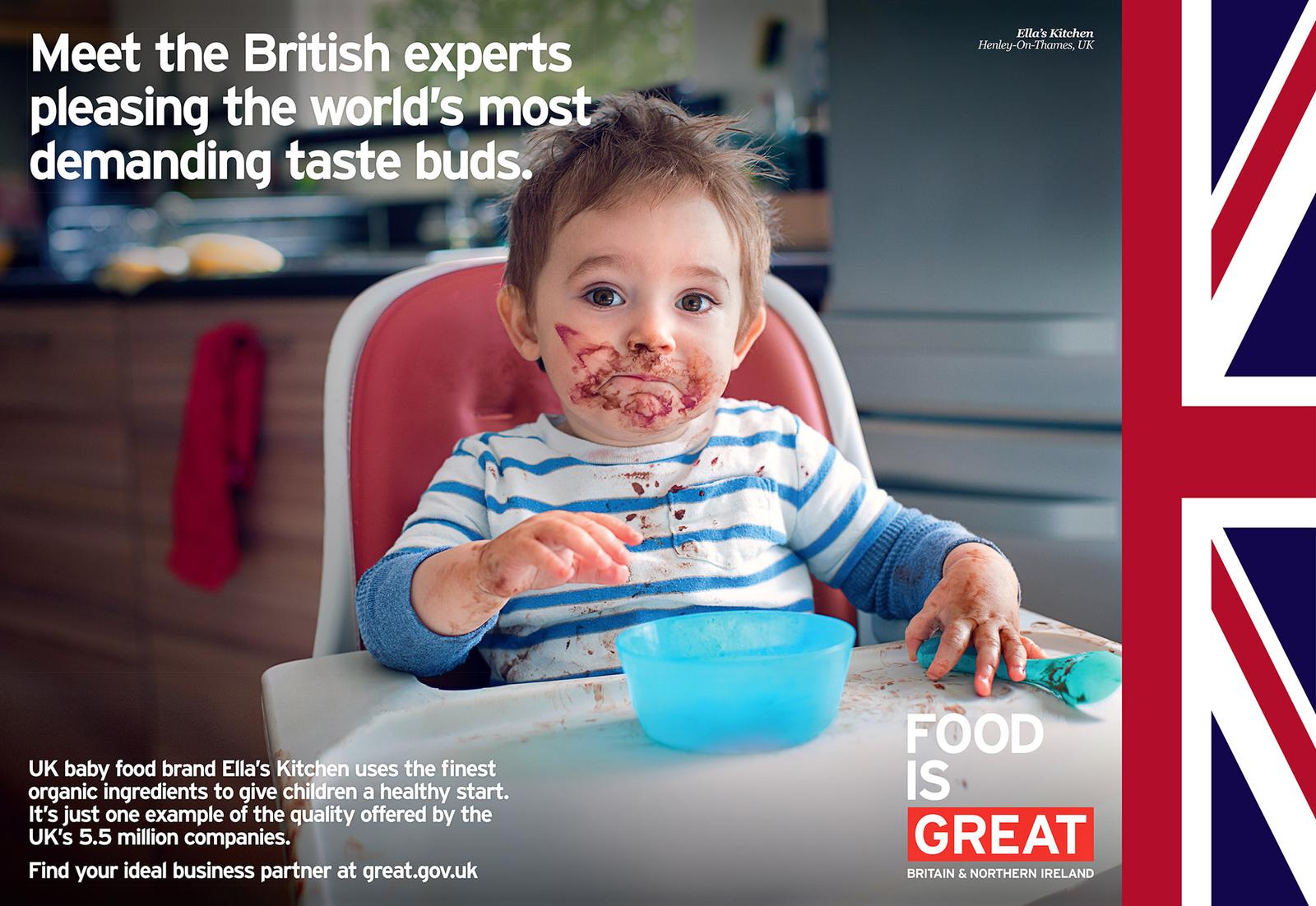 A toddler sits in a high chair, covered in chocolate and food, with a messy face and a spoon in hand, in a kitchen.