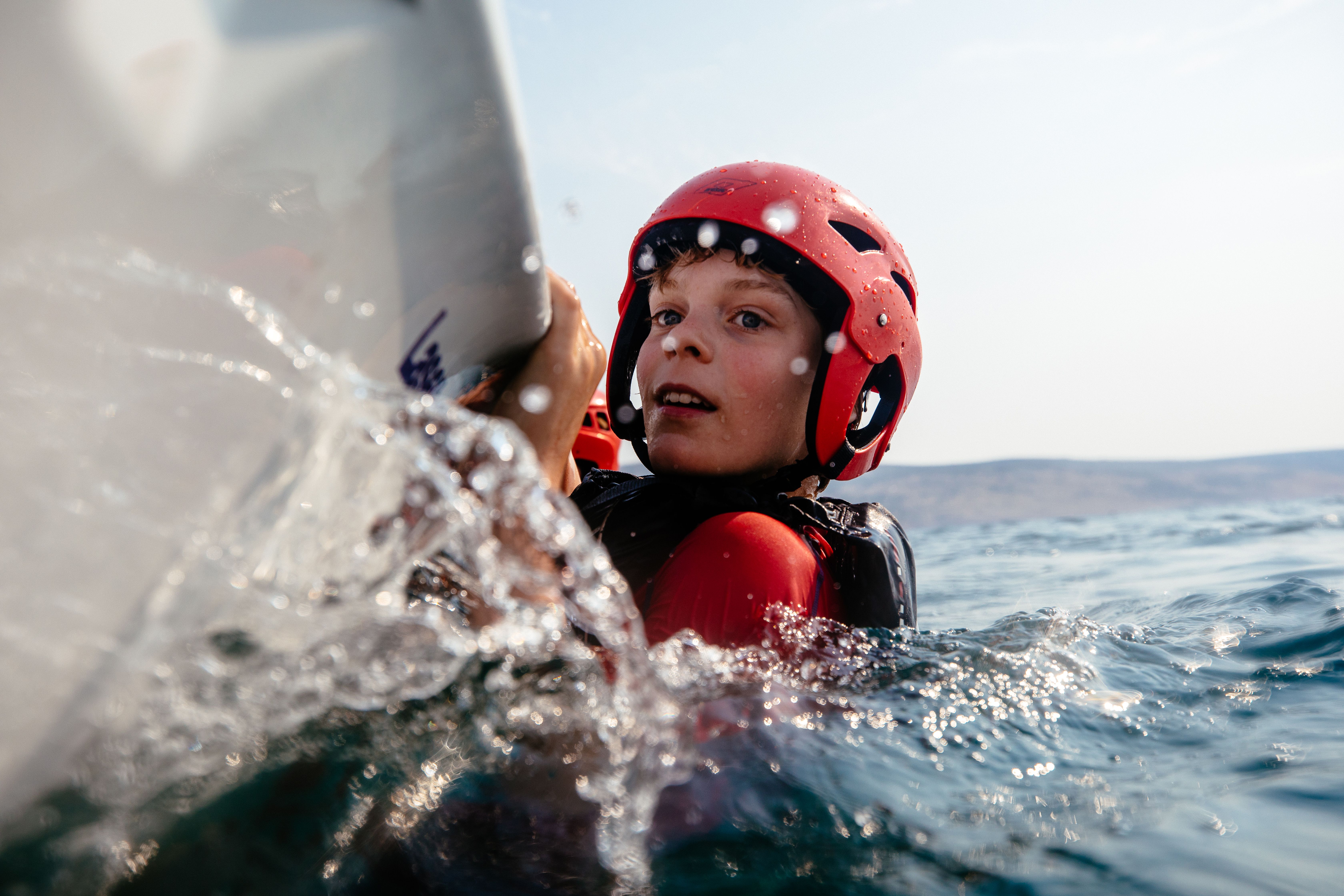 A young boy in a red wetsuit and helmet is holding onto a surfboard in the ocean, with a wave crashing in the background.