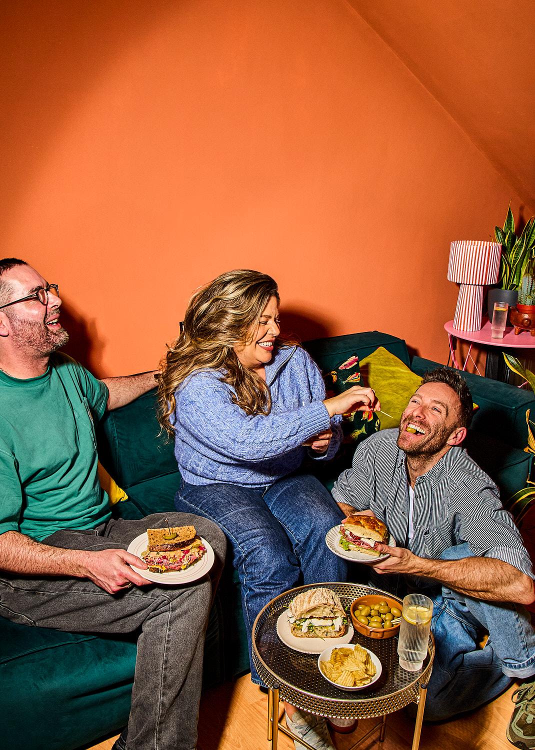 The image shows three friends laughing and enjoying a meal together on a dark green velvet couch. They are surrounded by various sandwiches, olives, and crisps on a small gold table, captured in Noemi Scavo’s signature bright, high-contrast style against an orange wall.