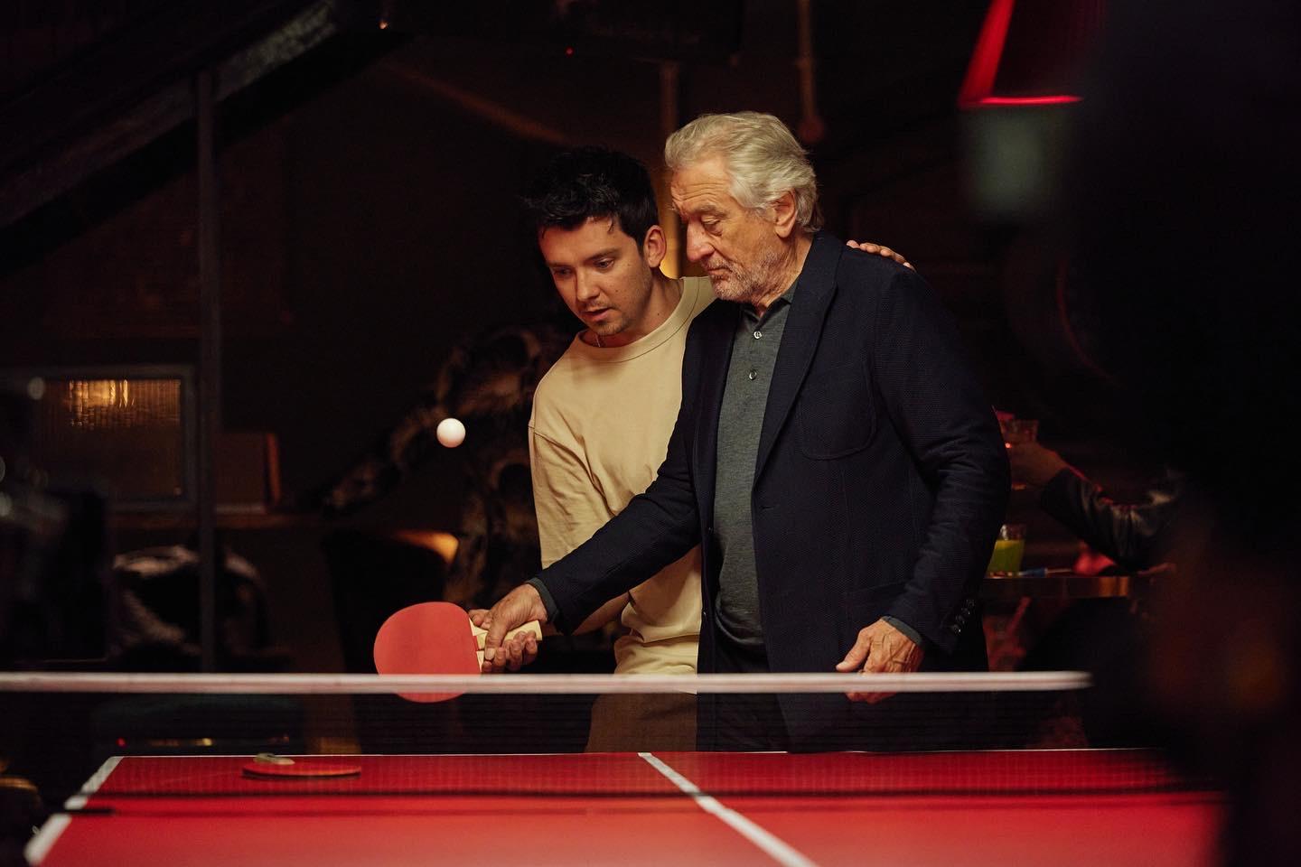 Two men, one older and one younger, are standing behind a table tennis table, with the younger man holding a table tennis paddle and the older man placing his hand on his shoulder.