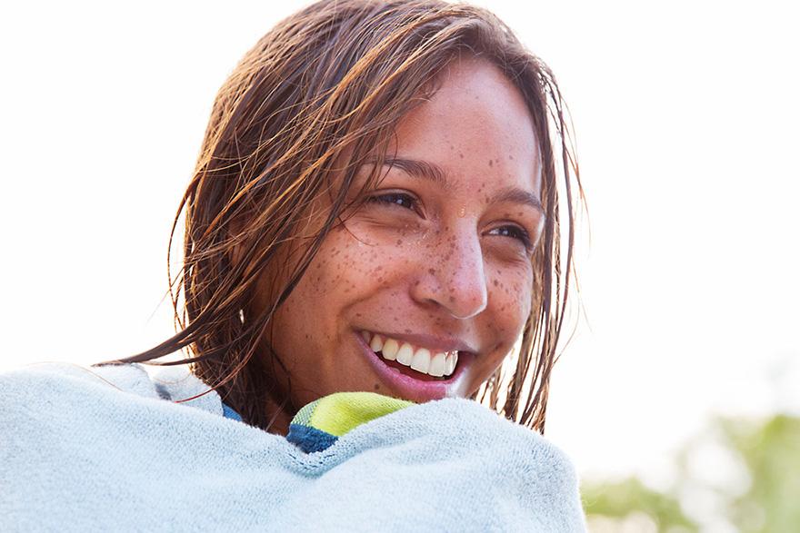 A young woman with long brown hair and freckles, smiling and wrapped in a white towel, with a beach or pool environment in the background.