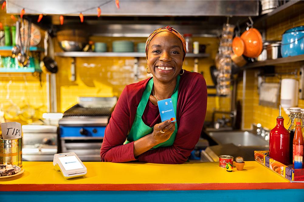 A woman stands behind a counter in a kitchen holding a blue card.
