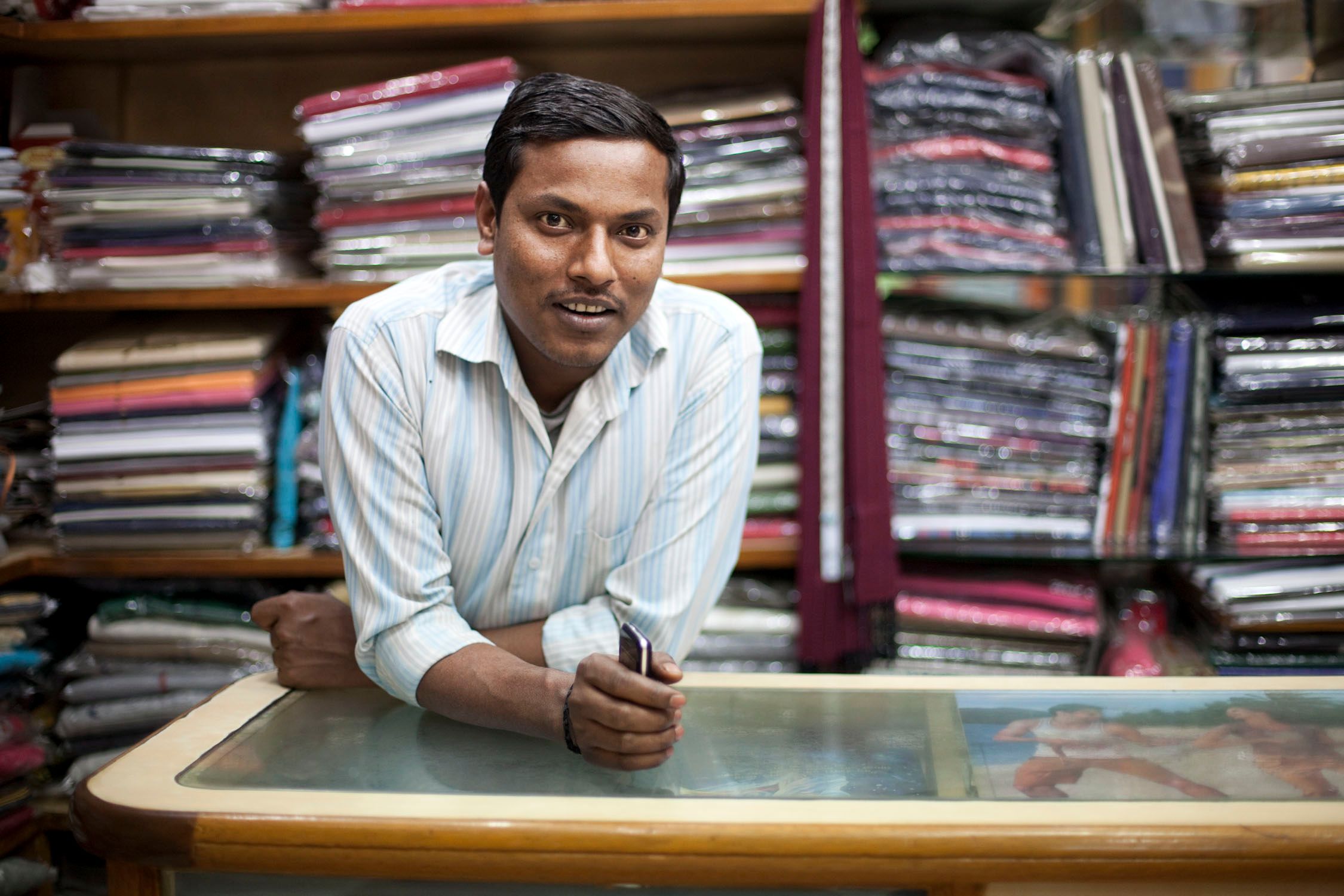 A man in a white and blue striped shirt leaning on a glass-topped table in front of a wall of fabric rolls.