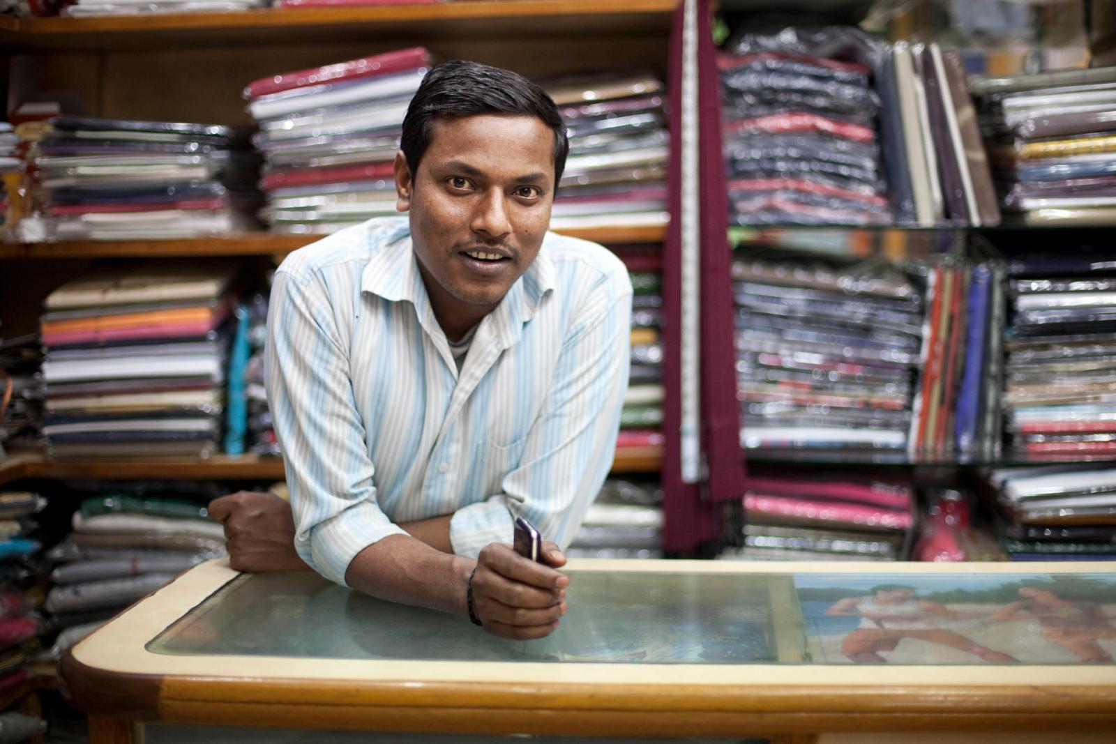 A man in a white and blue striped shirt leaning on a glass-topped table in front of a wall of fabric rolls.