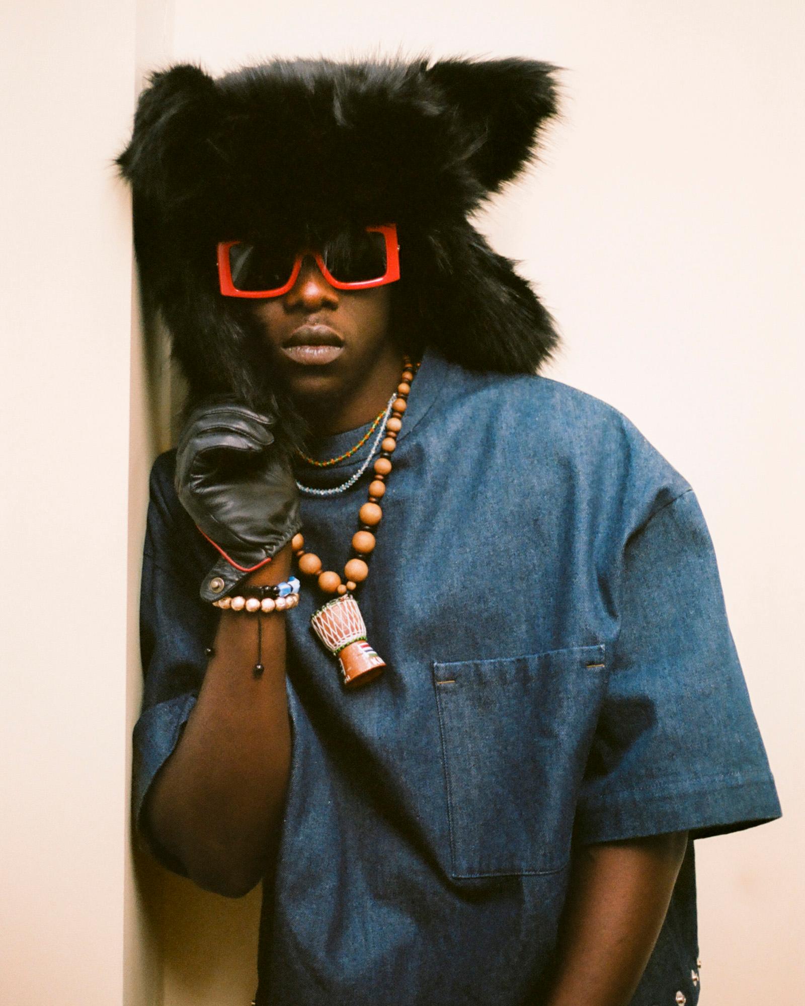 Kemka Ajoku wearing a black fur-trimmed hat, a denim jacket, and a gold chain, with a large, ornate, African-style necklace around his neck, and a pair of sunglasses on his face.