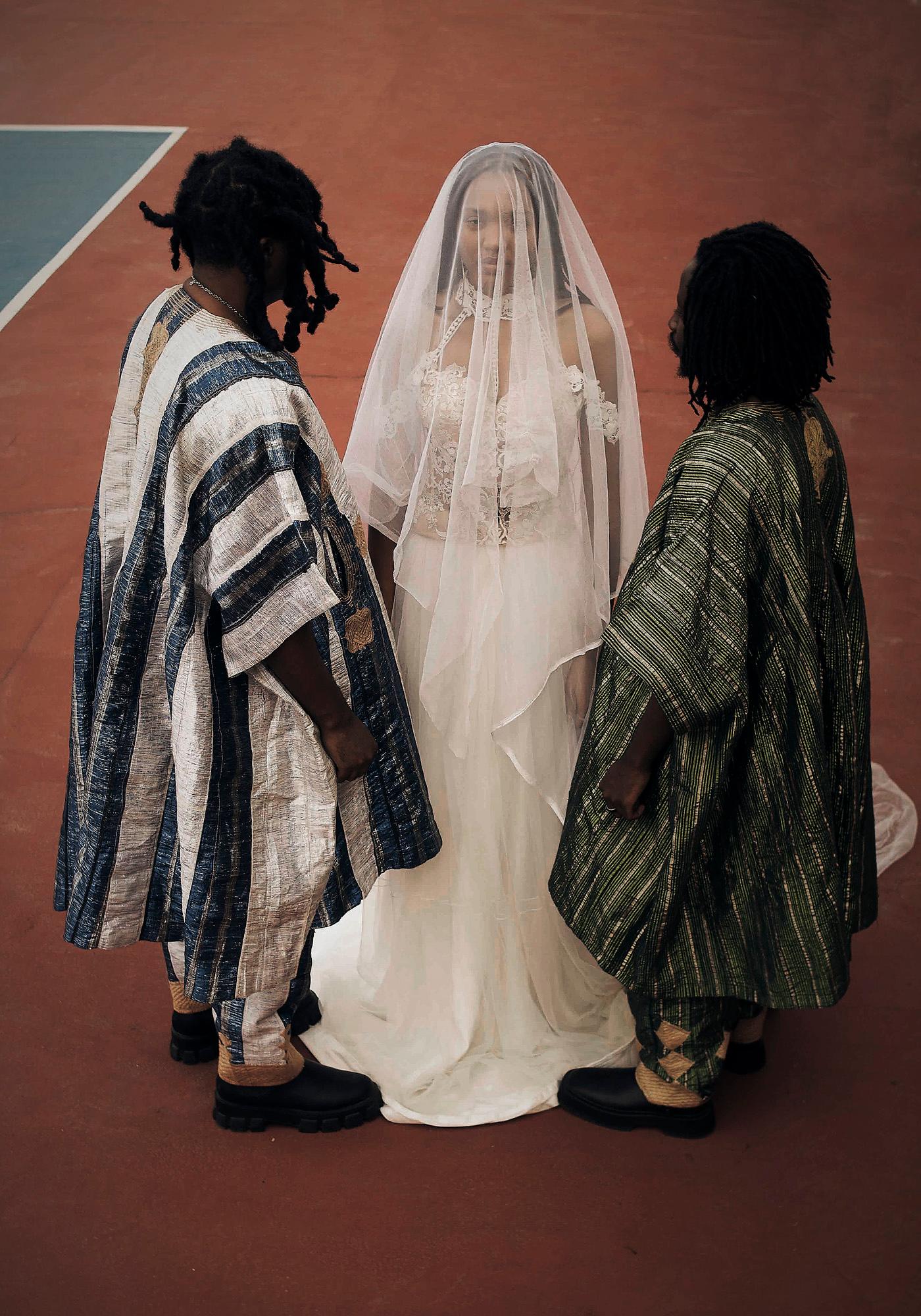 A bride in a white wedding dress with two men in traditional African attire nearby.
