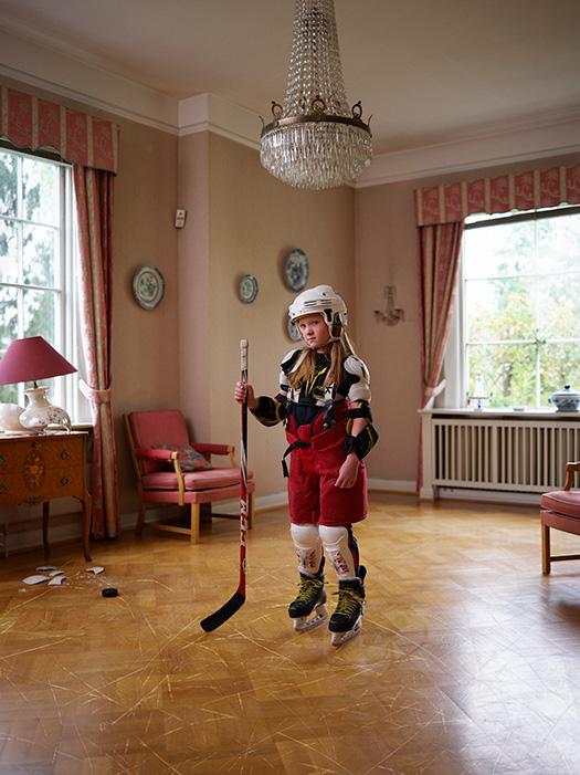 A young girl in a hockey outfit, holding a hockey stick, in a living room with a chandelier, a chair, a table, and a window.