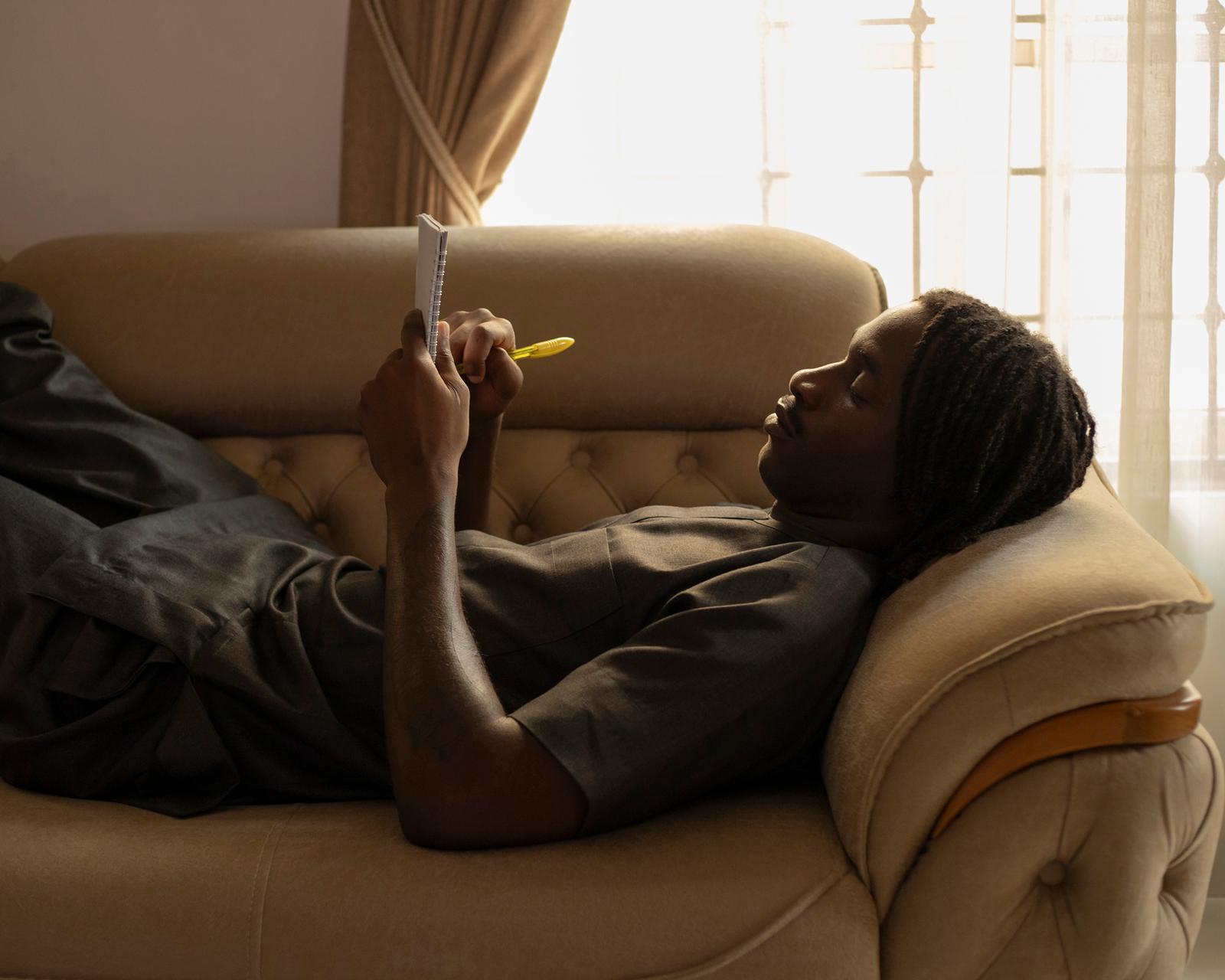 A person, likely the artist, is lying on a couch, holding a notebook and pen, and appears to be deep in thought.