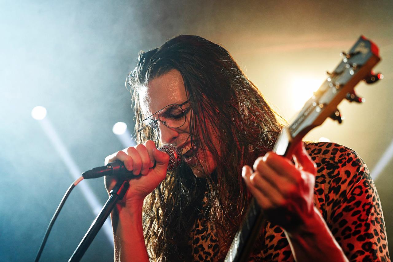 A man with long, wet hair and a leopard-print shirt is singing into a microphone while holding a guitar.