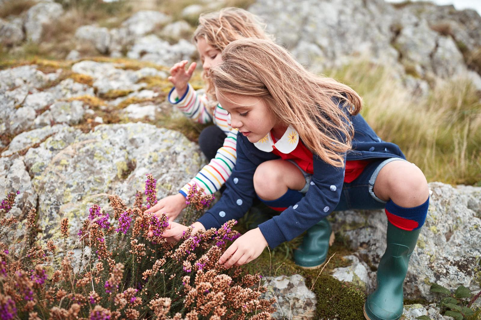 A young girl with long blonde hair kneels among wildflowers, her eyes fixed on a patch of purple heather. Her face is intent, her fingers reaching out to touch the delicate.