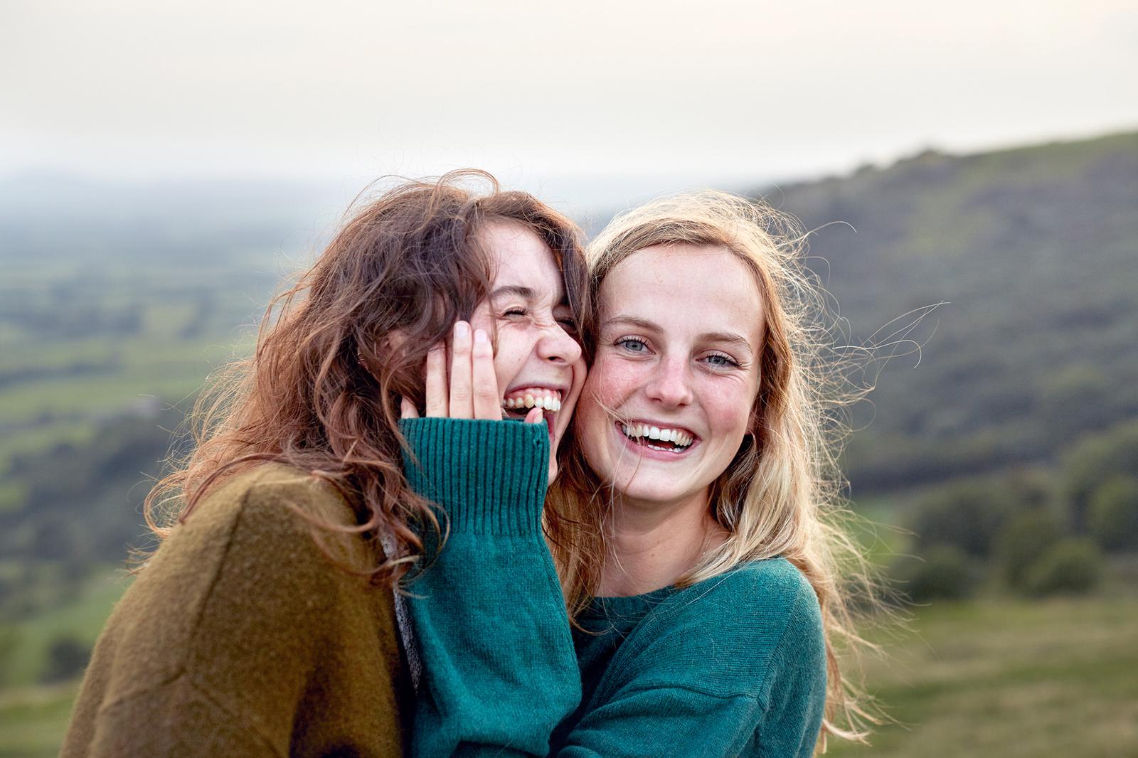 Two young women with long hair, smiling and embracing each other, both wearing green sweaters.