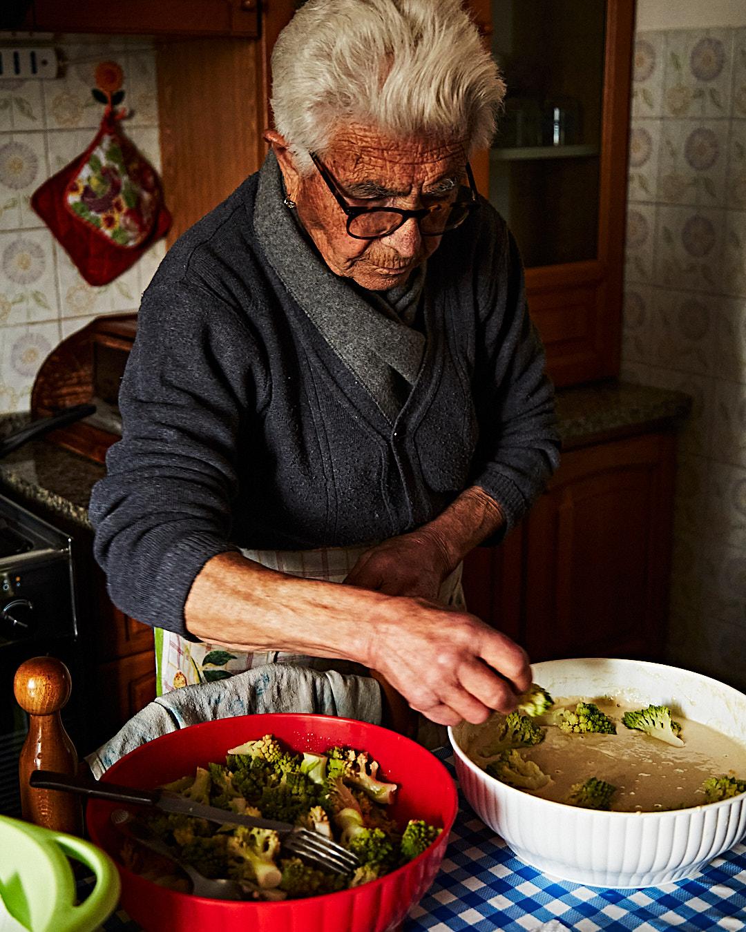An elderly woman with short white hair, wearing a dark grey cardigan over a grey scarf, preparing a meal in a kitchen.