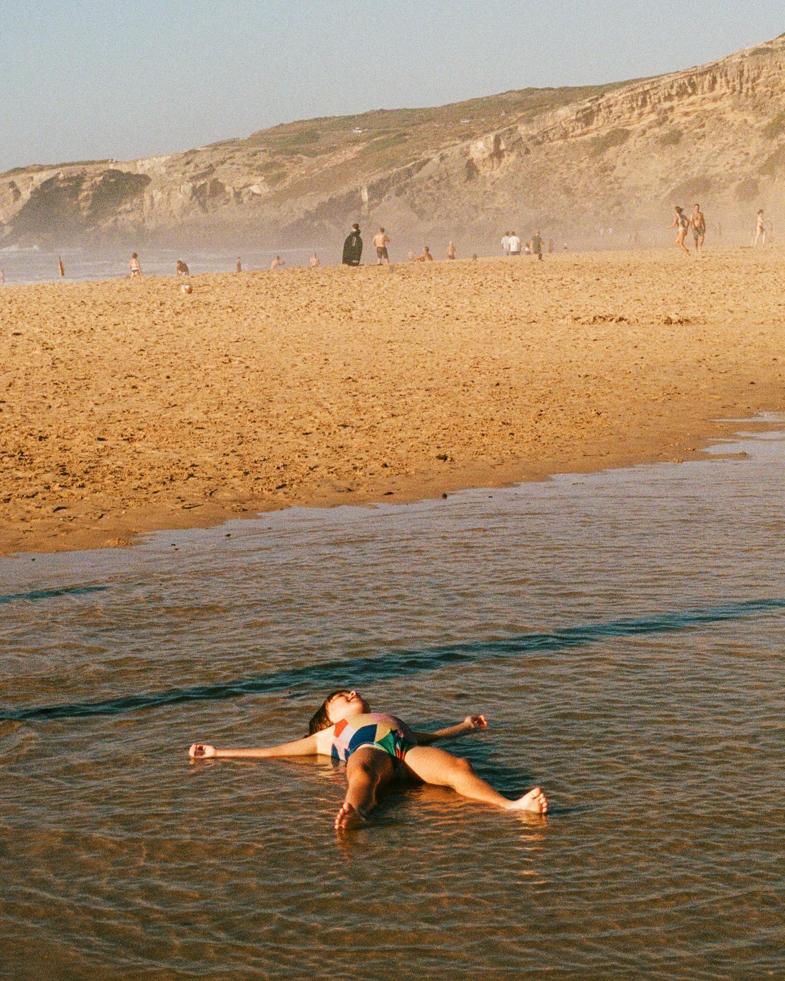 A woman in a swimsuit floating on her back in shallow water with her limbs relaxed and eyes closed.