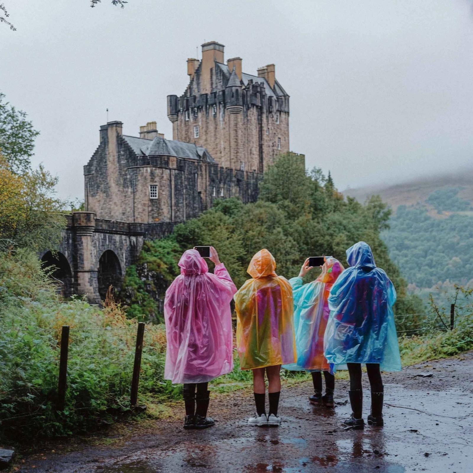 A group of people, likely tourists, wearing colorful rain ponchos, taking photos in front of a historic, stone castle in a lush, green, mountainous landscape.