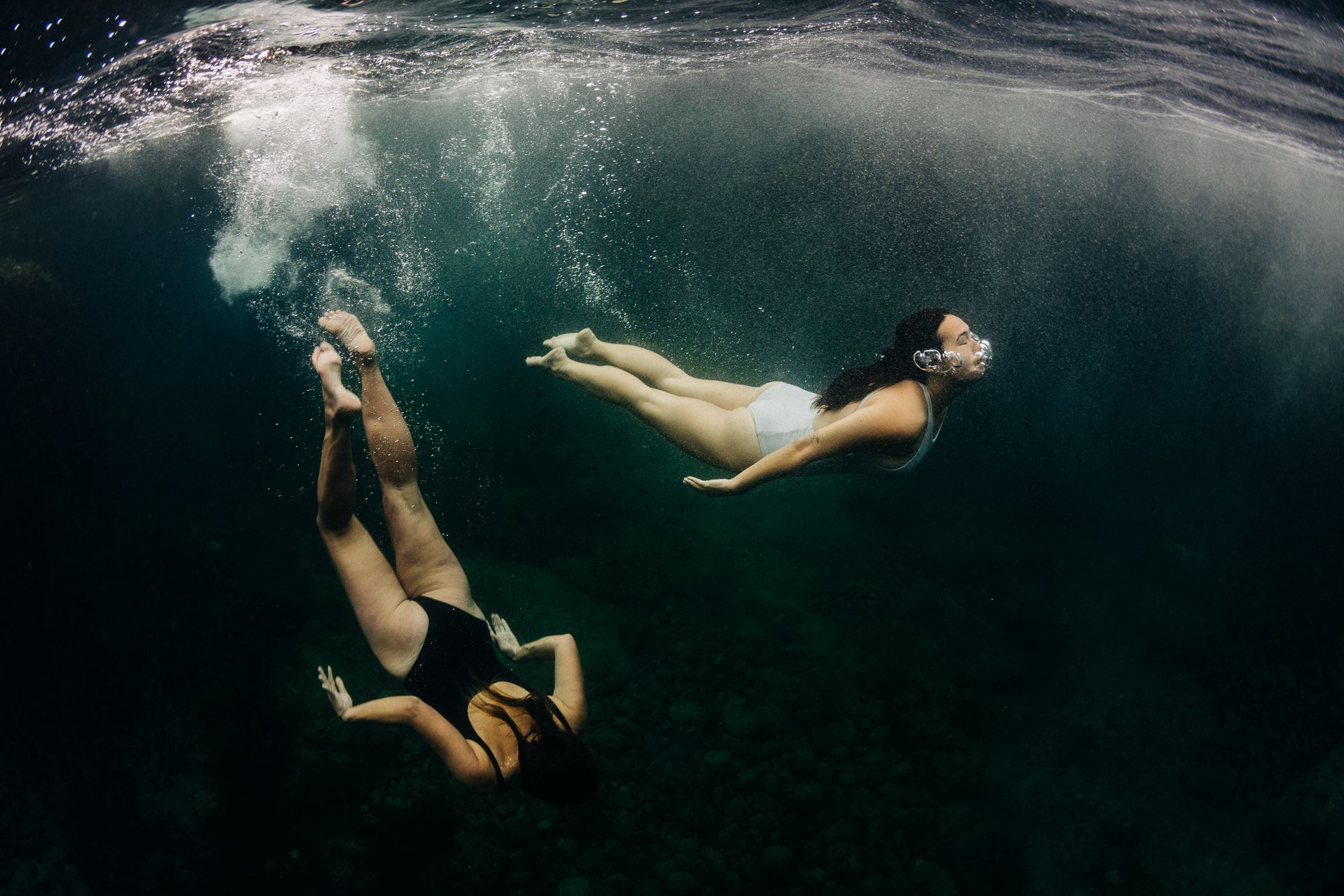 Two women in swimsuits and snorkeling masks are swimming underwater, surrounded by a school of fish.