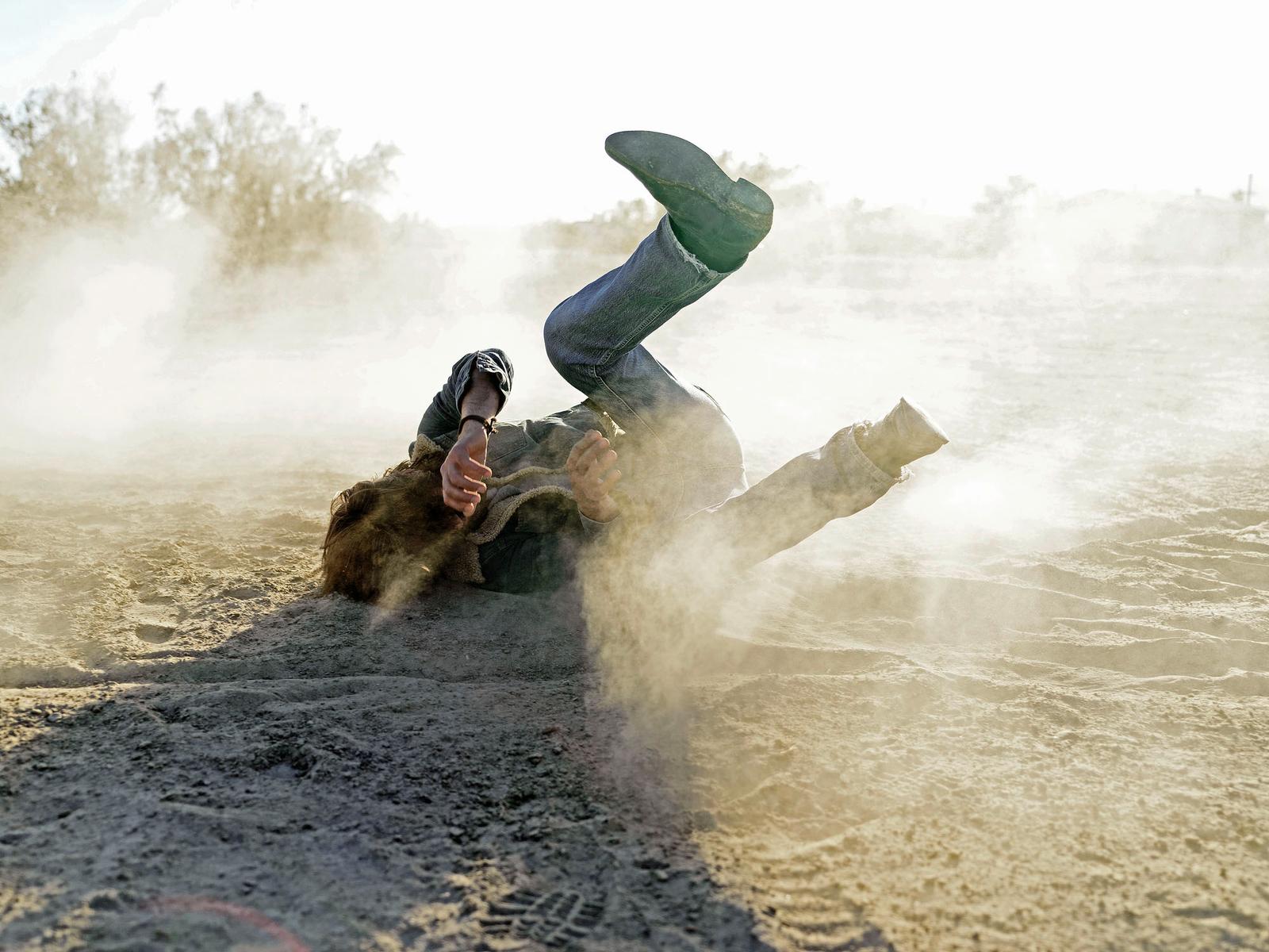 A man is buried in the sand, with his legs in the air, wearing a pair of jeans and a t-shirt, in a desert-like environment.