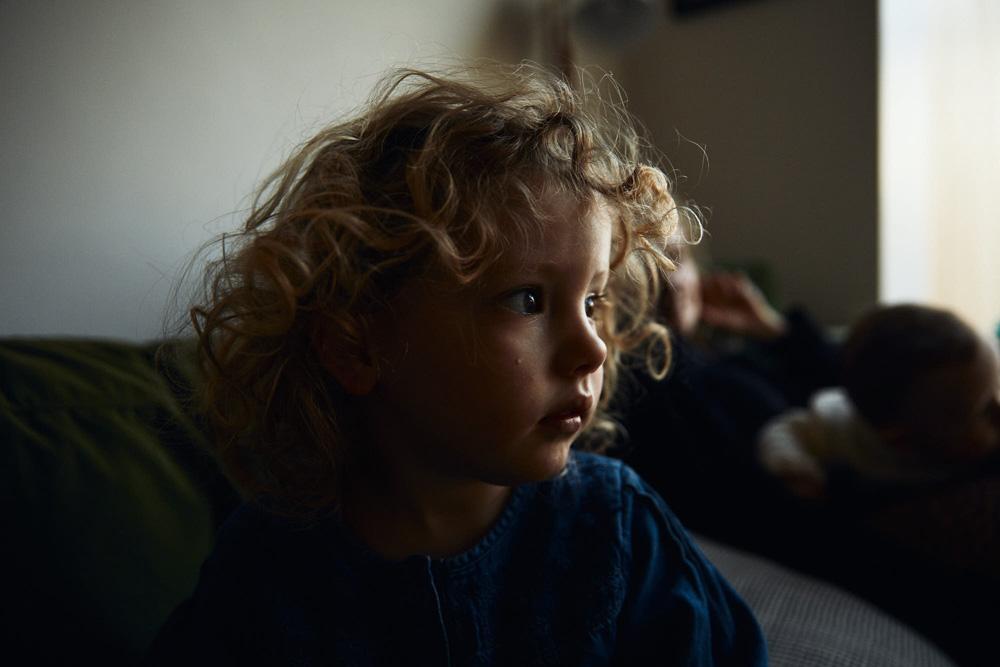 A young child with curly blonde hair and a blue shirt sits on a couch, looking up at something with a mix of curiosity and concern on their face.