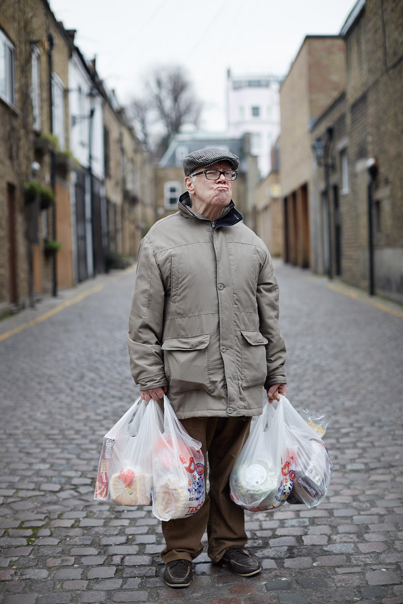 An elderly man in a grey coat, brown trousers, and a flat cap, carrying multiple plastic shopping bags down a narrow, cobblestone street lined with old, brick buildings.