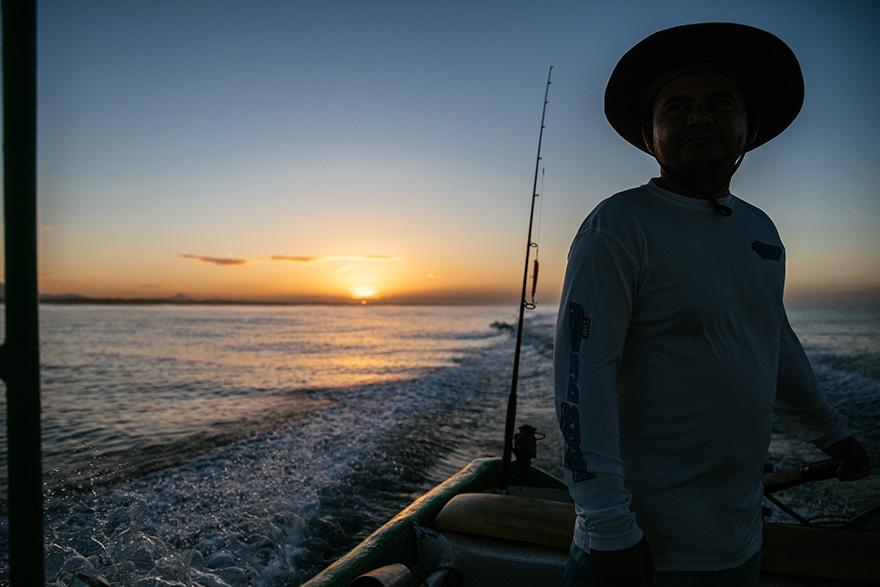 A man in a wide-brimmed hat and a long-sleeved shirt is standing on a boat in the water, with a fishing rod in the background.