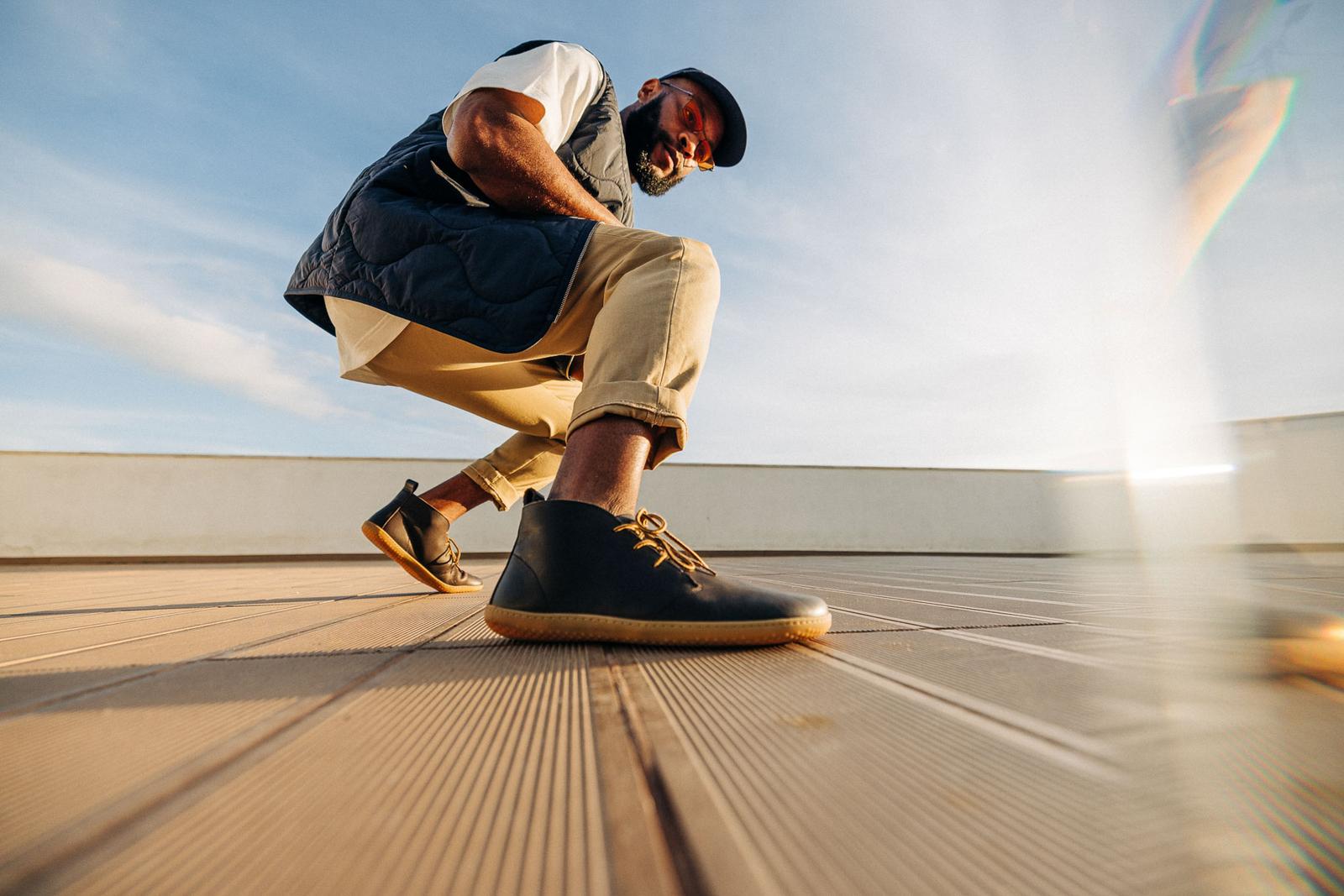 A man in a black and white outfit, wearing a black puffer vest, white t-shirt, and beige trousers, posing on a wooden floor, with a blue sky and white clouds in the background.
