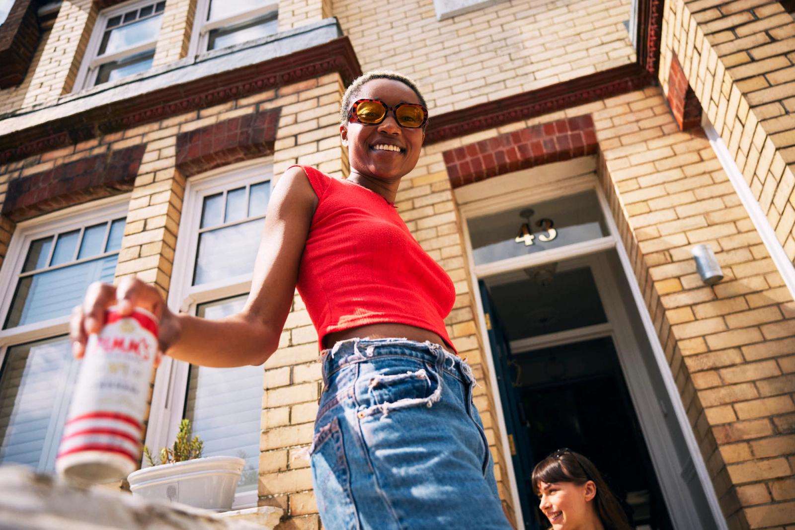 A woman in a red top and blue jeans stands in front of a brick building, holding a can of beer and smiling at the camera. The woman is wearing sunglasses and has a relaxed, happy.
