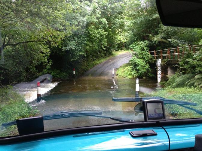 A vehicle is driving through a flooded road in a forest, with the water level rising to the vehicle's wheels.