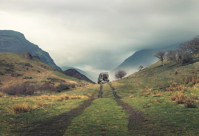 A 4x4 vehicle driving down a dirt road in a mountainous landscape, with a rugged hillside to the left and a valley to the right.