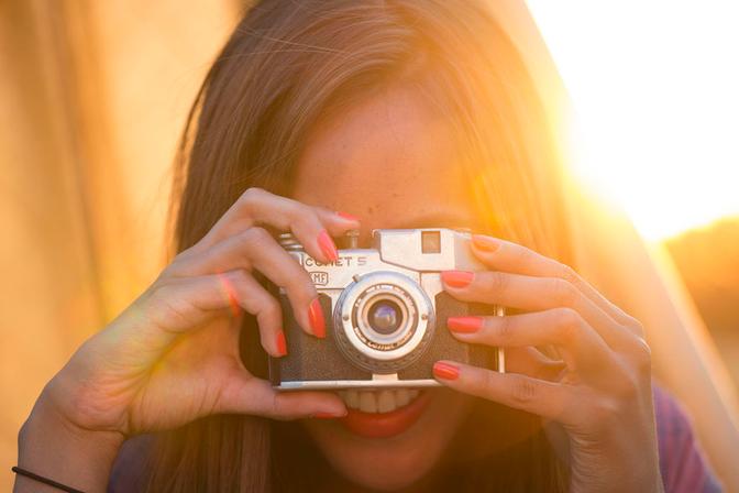A woman holding a vintage camera, smiling and looking through the viewfinder.