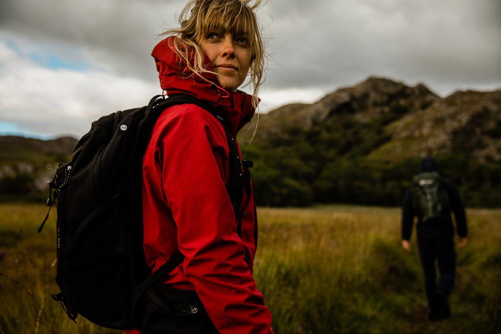 A woman in a red jacket and backpack stands in a mountainous landscape with a distant man.