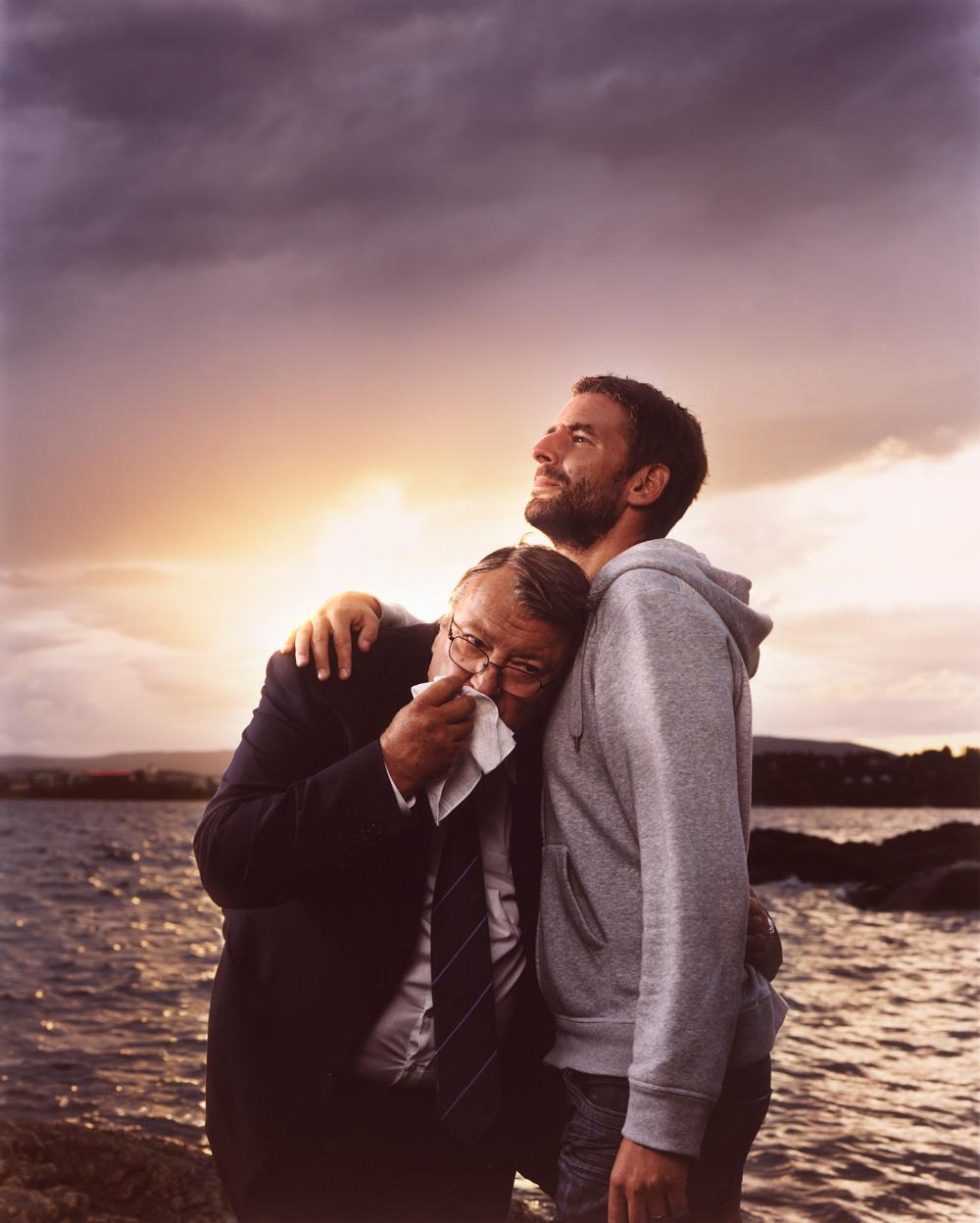 Two men, one in a suit and one in a hoodie, embracing by the water at sunset.