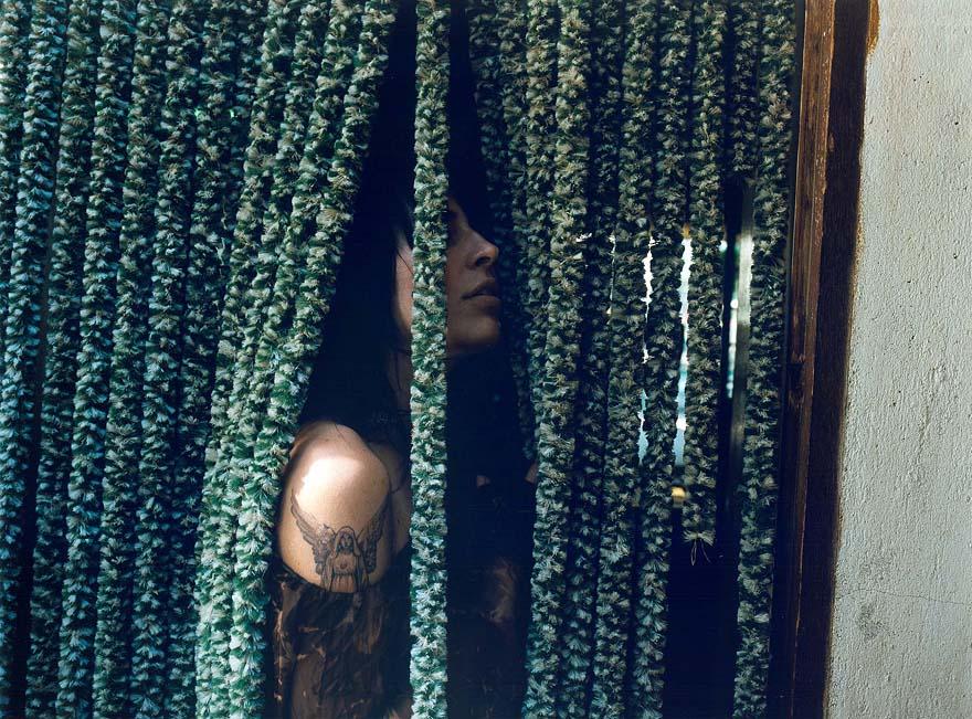 A person with tattoos is hiding behind a curtain of green, fuzzy, textured strands.
