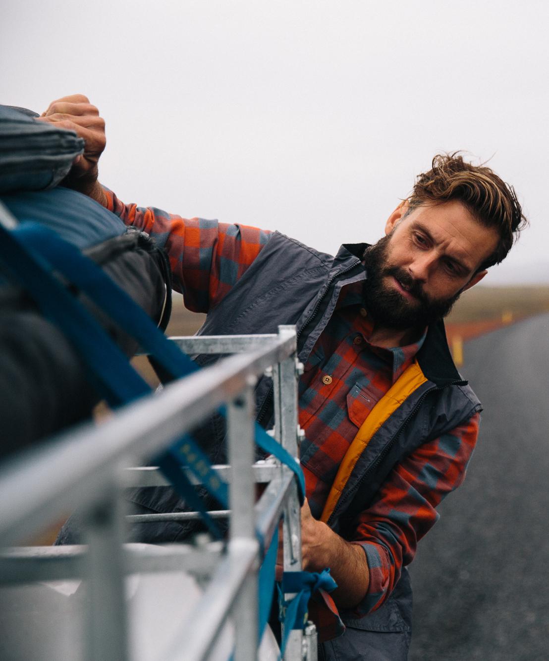 A man in a red and blue plaid shirt and a black jacket is standing on a road, holding a backpack and looking at the camera with a serious expression.