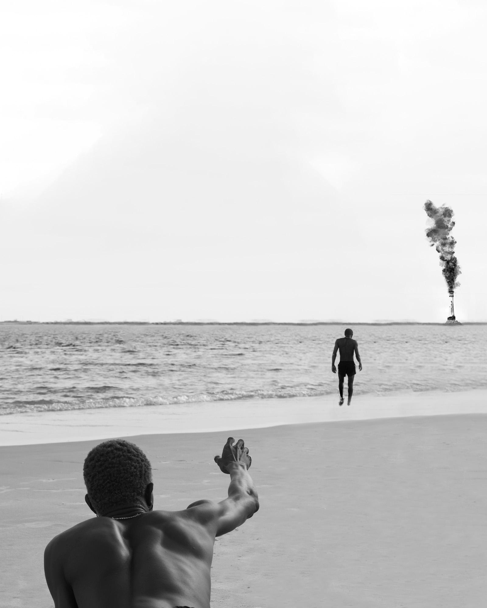 Two men on a beach, one in the foreground is waving at the other who is walking towards him, with a large plume of smoke in the background.