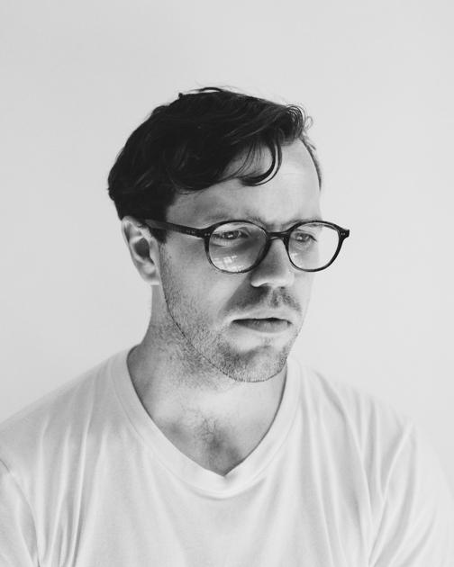 A man with a beard and glasses, wearing a white shirt and a black jacket, looking directly at the camera with a serious expression.